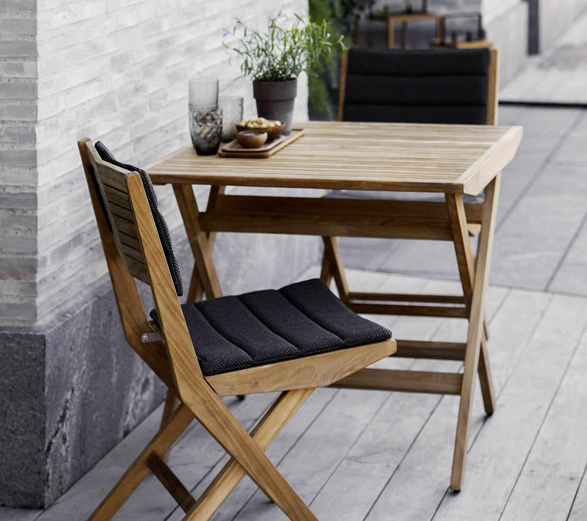 Houten tafel en stoelen set met zwarte kussens, versierd met potplanten en decoratief glaswerk. Perfect voor een gezellige setting.