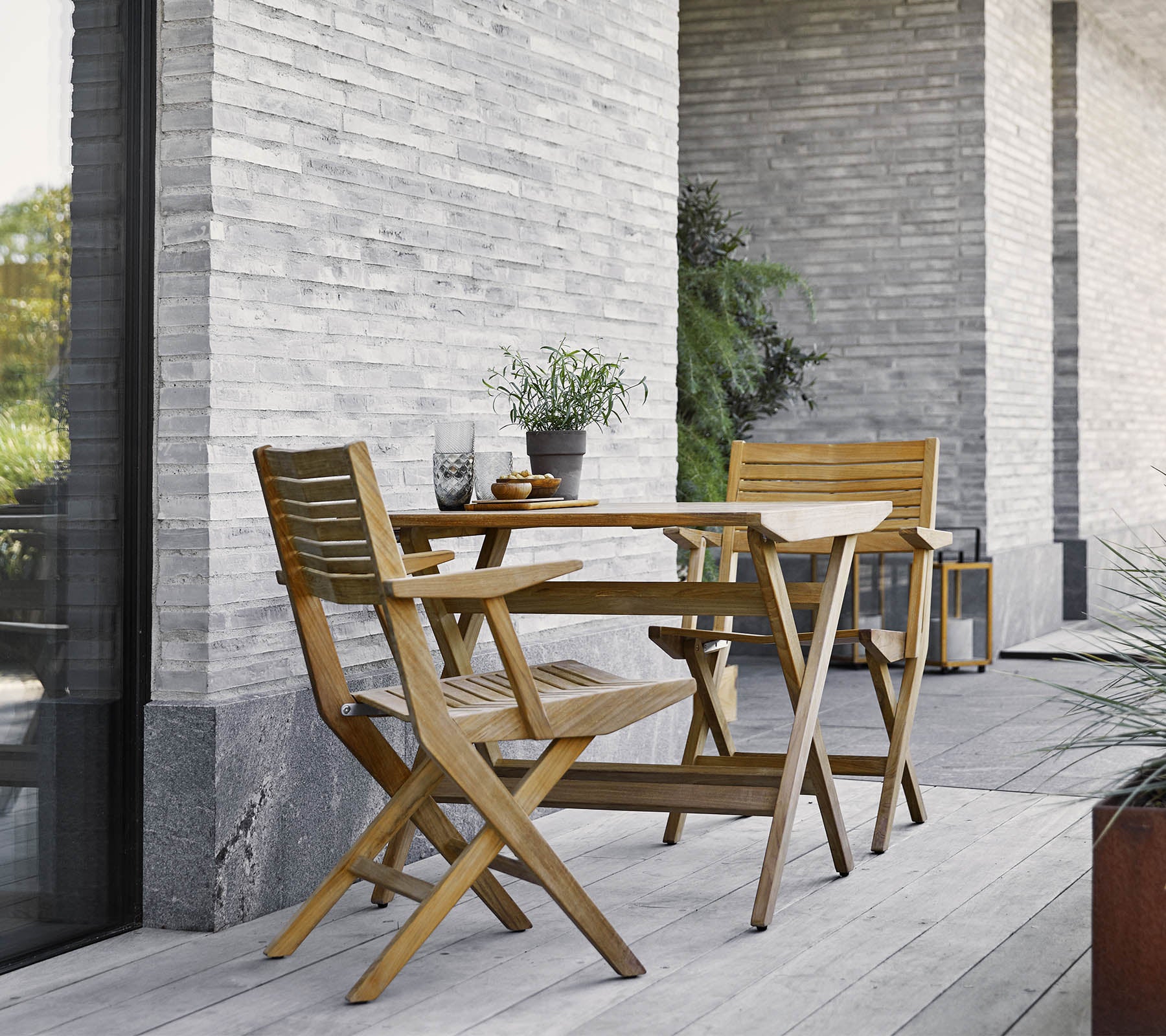 Een houten opklaptafel met twee bijpassende stoelen, versierd met potplanten, tegen een gestructureerde stenen muur.