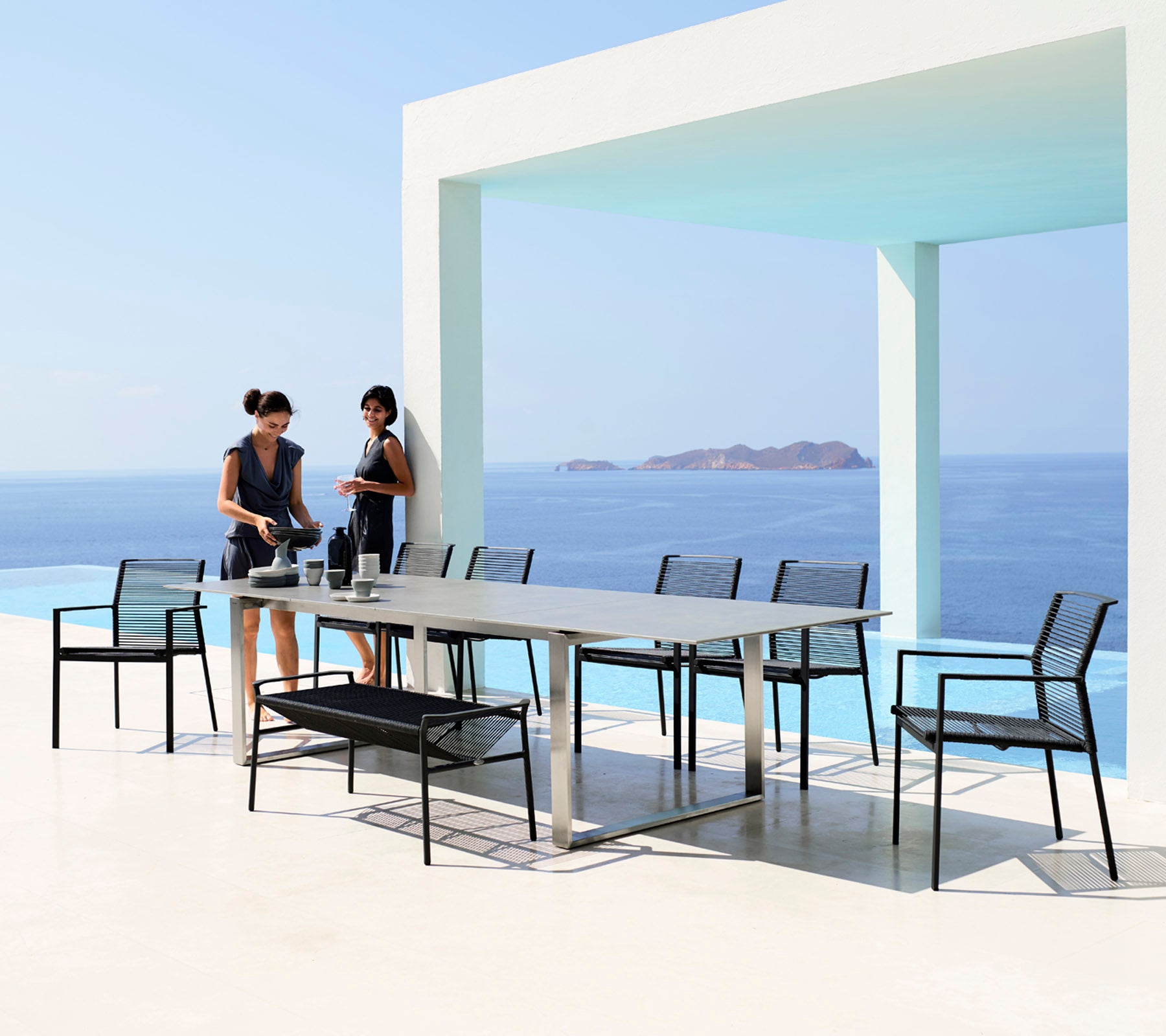 Twee vrouwen dekken een eettafel met elegante zwarte stoelen tegen de achtergrond van een serene blauwe zee en lucht.