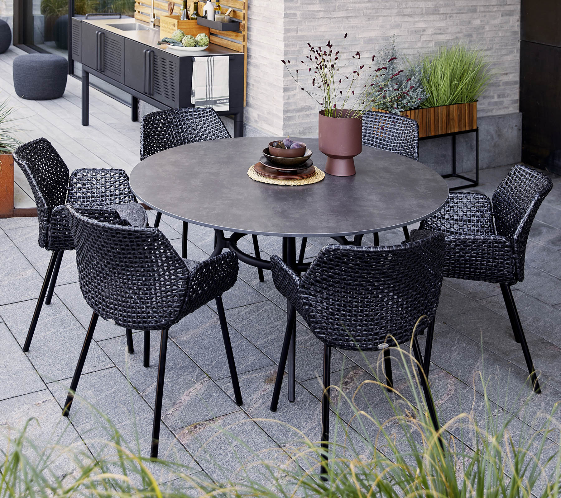 Ronde tafel met zes zwarte gevlochten stoelen, met een decoratieve schaal en een potplant, geplaatst tussen groen en moderne decoratie.