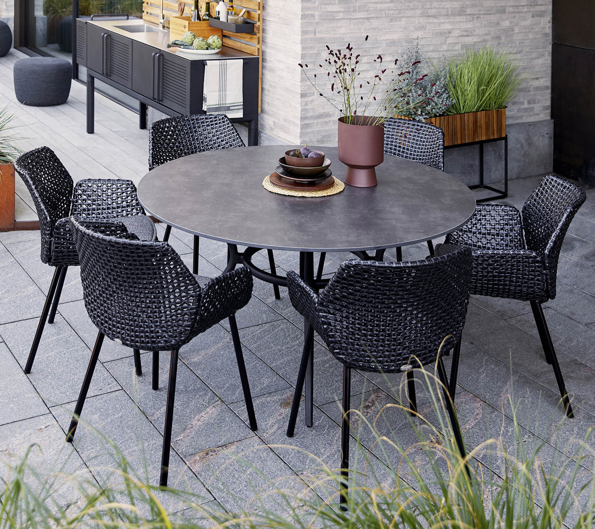 Ronde tafel met zes zwarte gevlochten stoelen, met een decoratieve schaal en een potplant, geplaatst tussen groen en moderne decoratie.