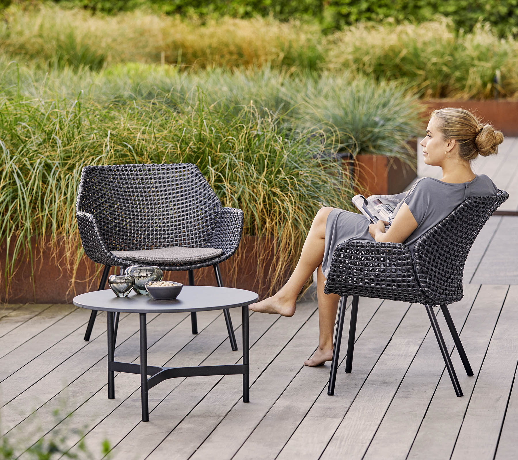Twee stijlvolle gevlochten stoelen met een bijpassende tafel, omringd door weelderig groen. Een vrouw ontspant terwijl ze een tijdschrift vasthoudt.
