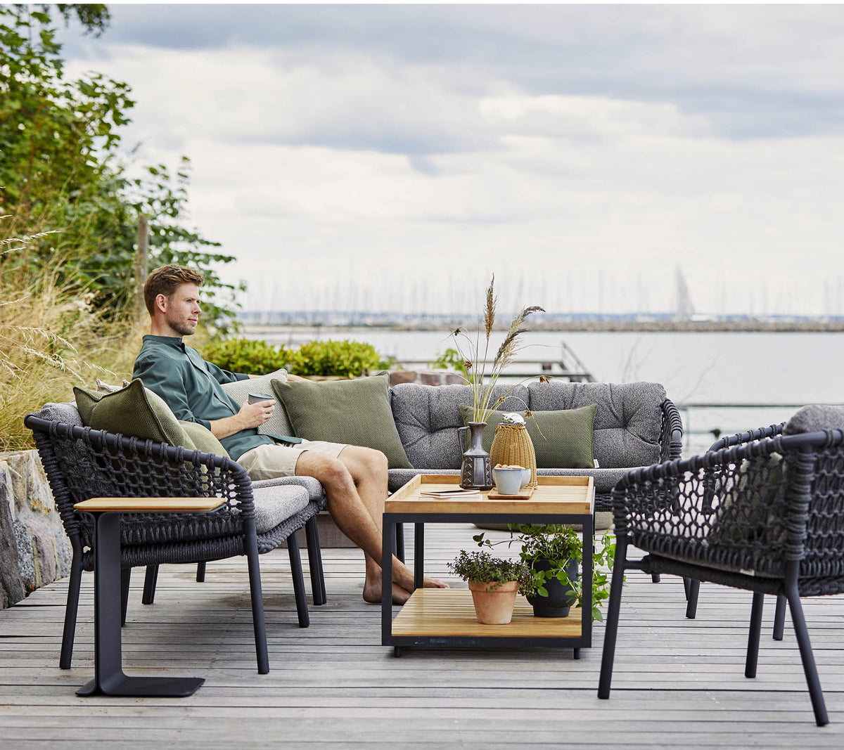 Een comfortabele zitruimte met een man die ontspant, met stijlvolle rieten stoelen en een houten salontafel versierd met planten en decoratieve items.