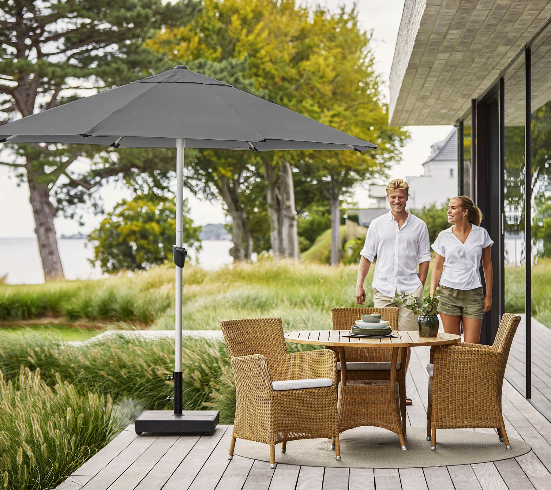 Een stel dat over een houten dek loopt met een ronde tafel en wicker stoelen, in de schaduw van een grote grijze parasol, met uitzicht op weelderig groen.
