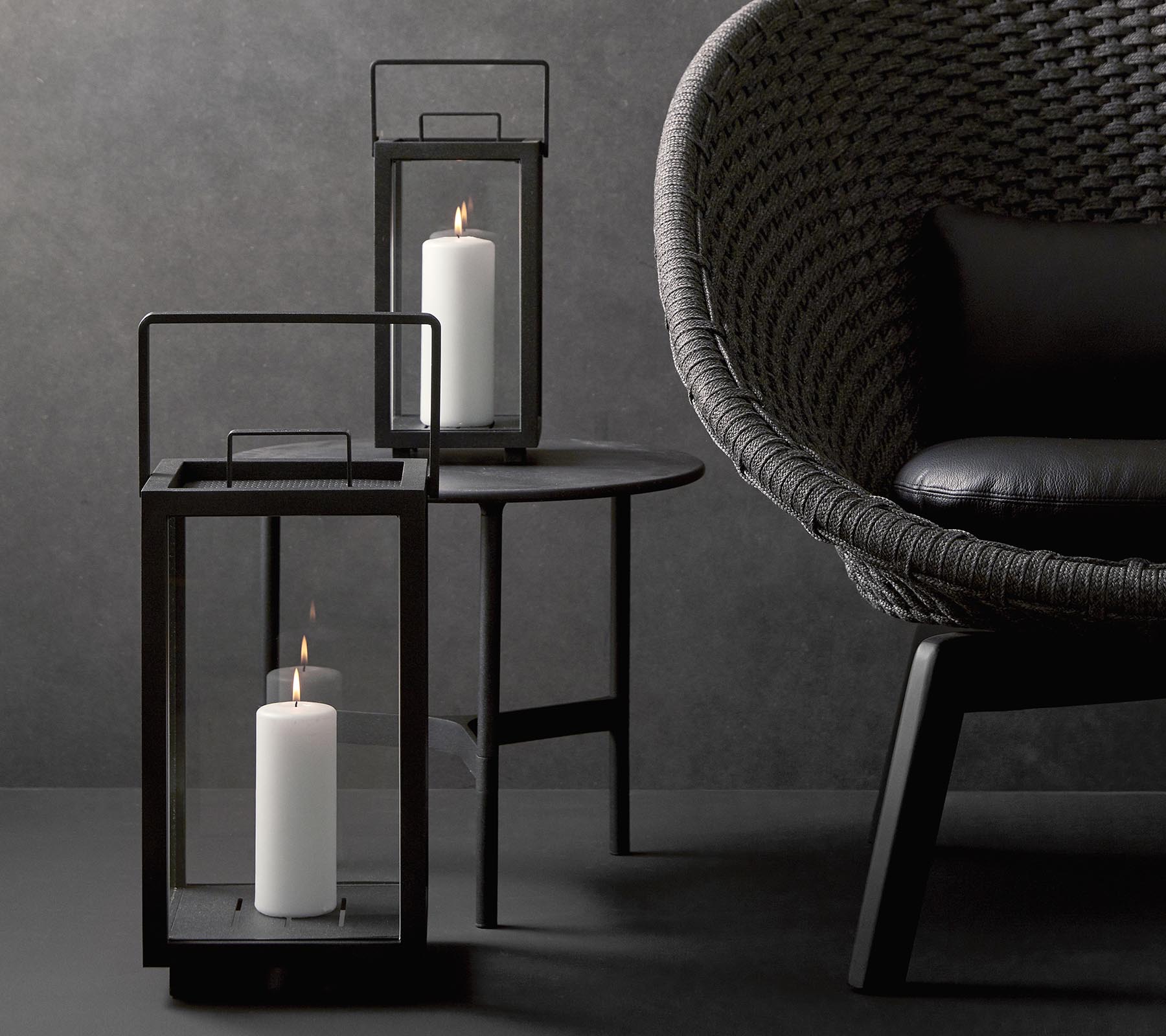 Twee stijlvolle lantaarns met witte kaarsen, vergezeld van een moderne, gestructureerde stoel aan een minimalistische tafel tegen een donkere achtergrond.