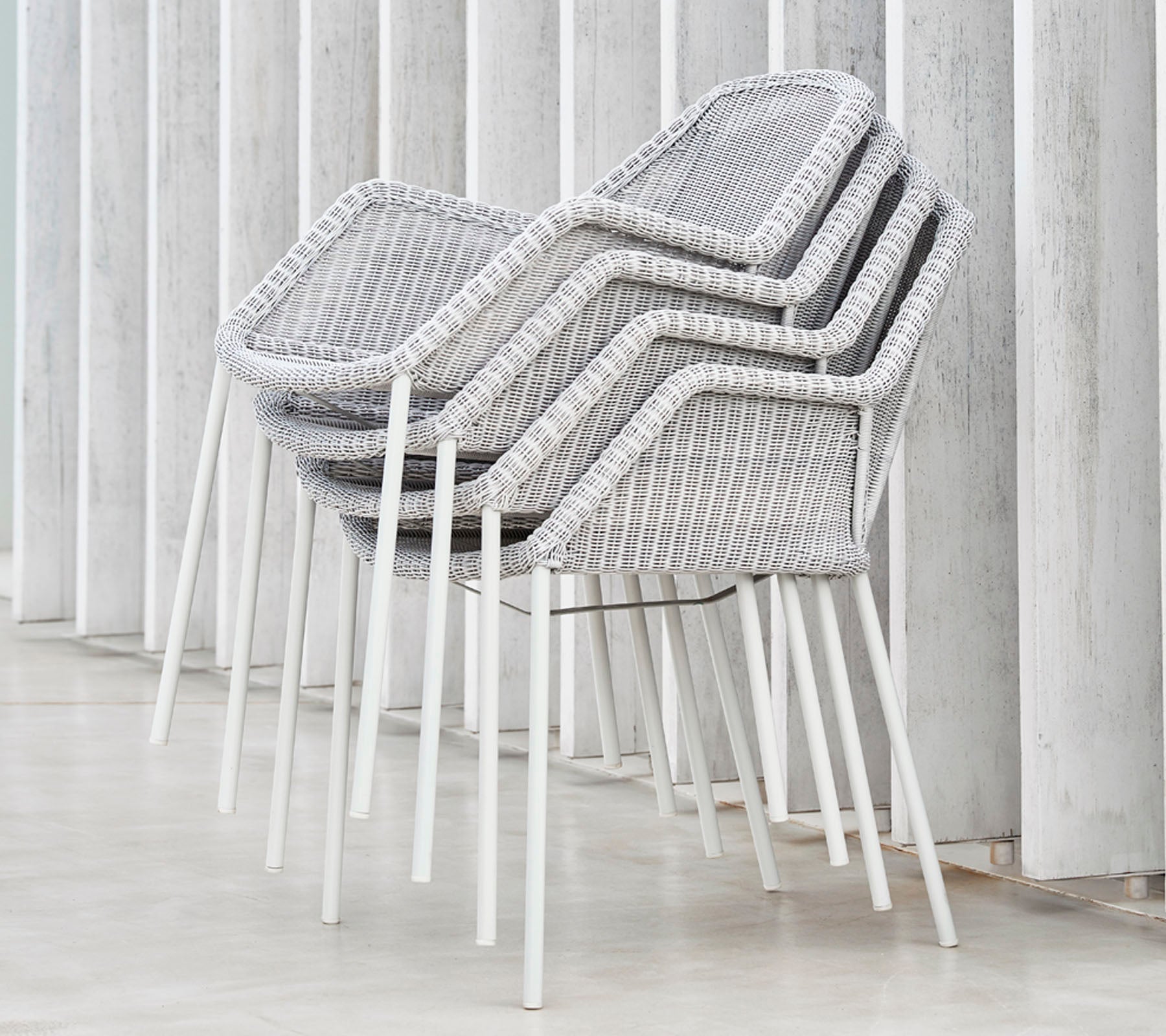 Gestapelde moderne stoelen met een geweven ontwerp in grijs en wit, leunend tegen verticale oppervlakken, en een strakke en minimalistische esthetiek tentoonstellend.