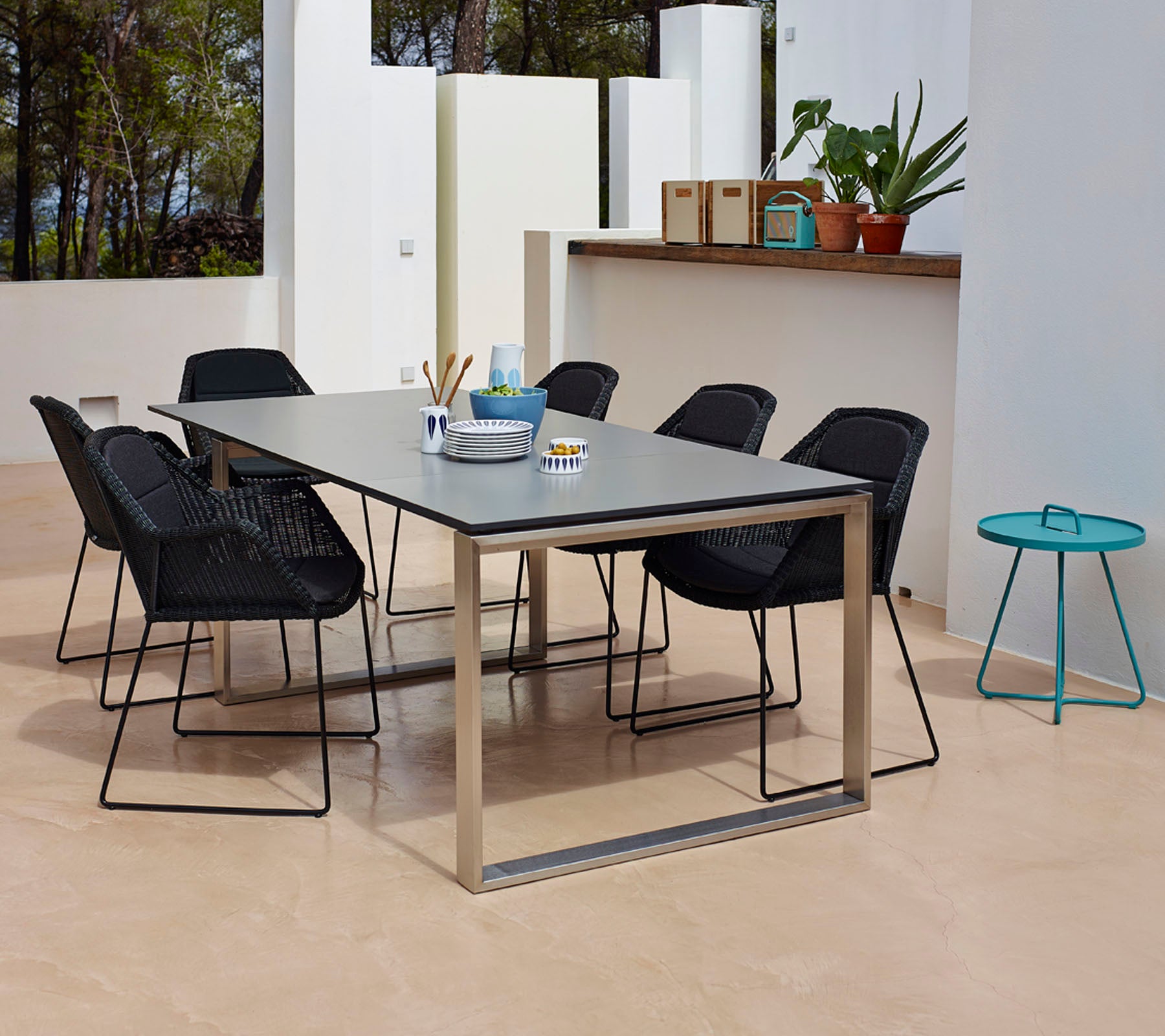 Een moderne eetopstelling met een rechthoekige tafel met een glad oppervlak, omringd door zes zwarte gevlochten stoelen en een klein teal bijzettafel.