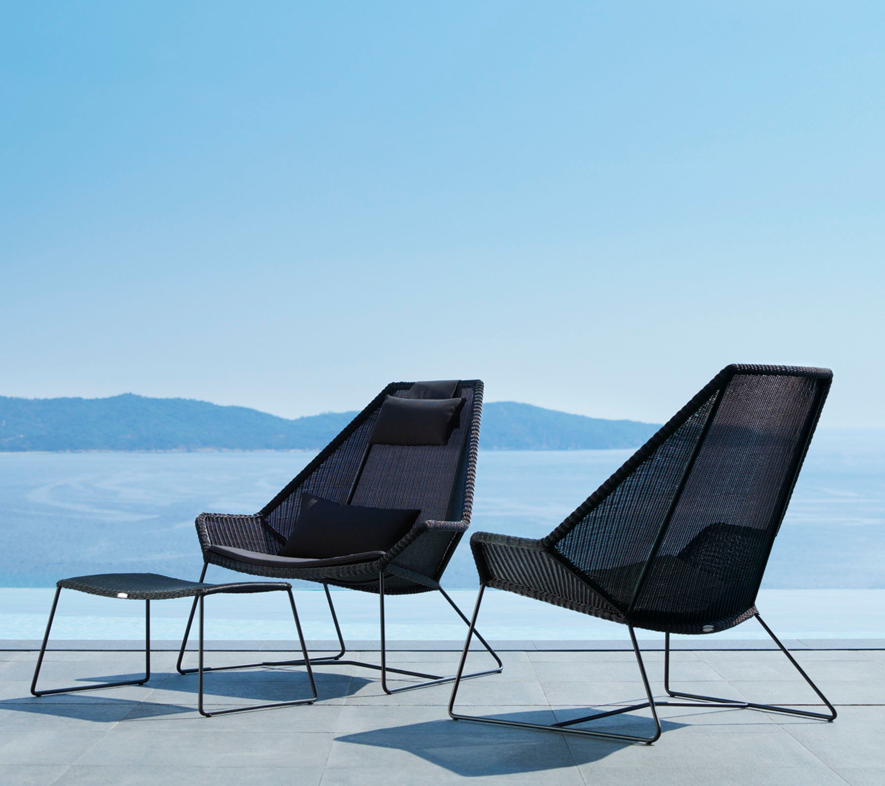 Twee stijlvolle, moderne stoelen met strakke lijnen en een open mesh-ontwerp, vergezeld van een bijpassende voetensteun, met uitzicht op een sereen waterlandschap.