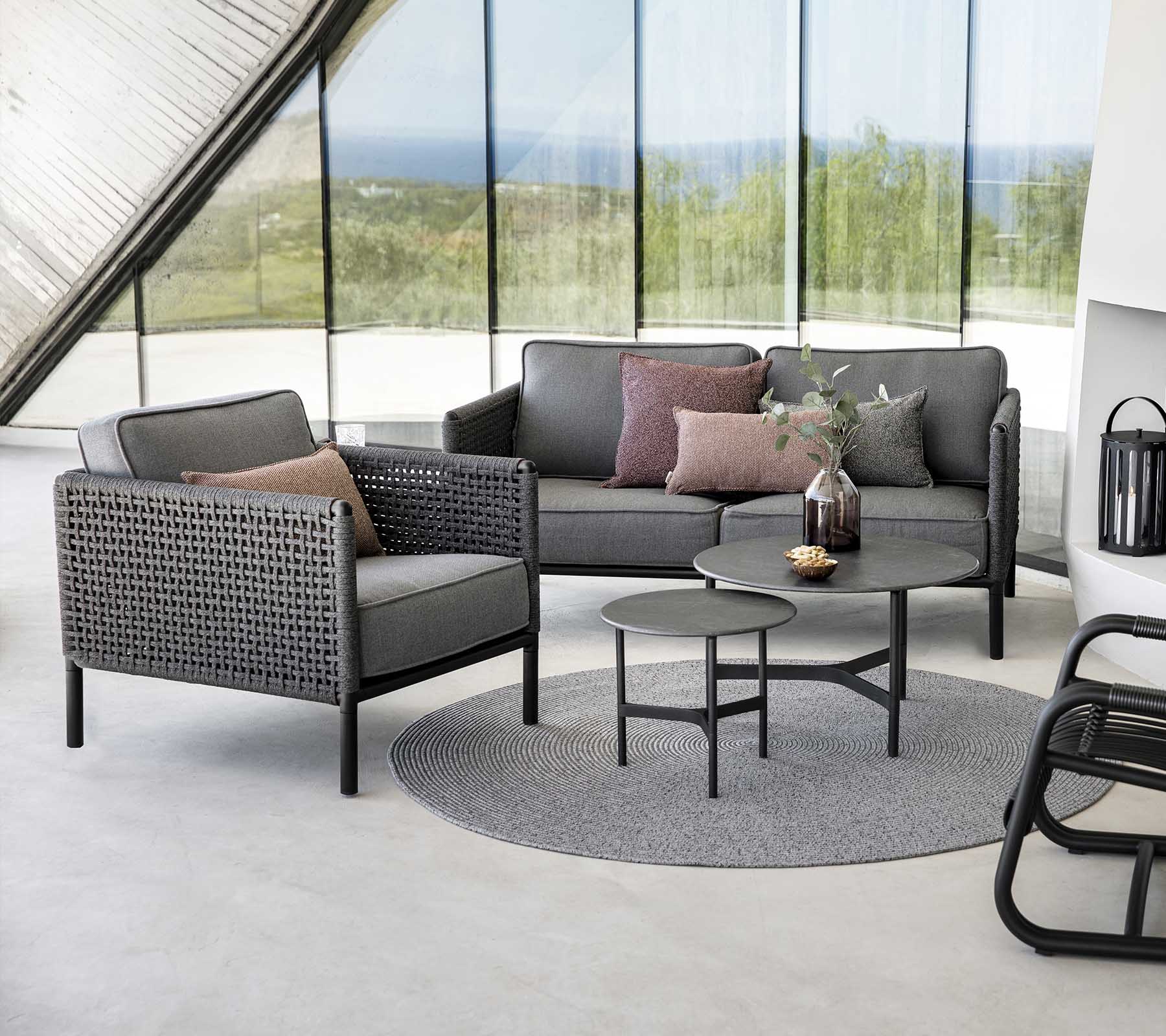 Stijlvolle zitopstelling met een grijze bank en fauteuil met kussens, vergezeld van een ronde tafel op een gestructureerd tapijt.