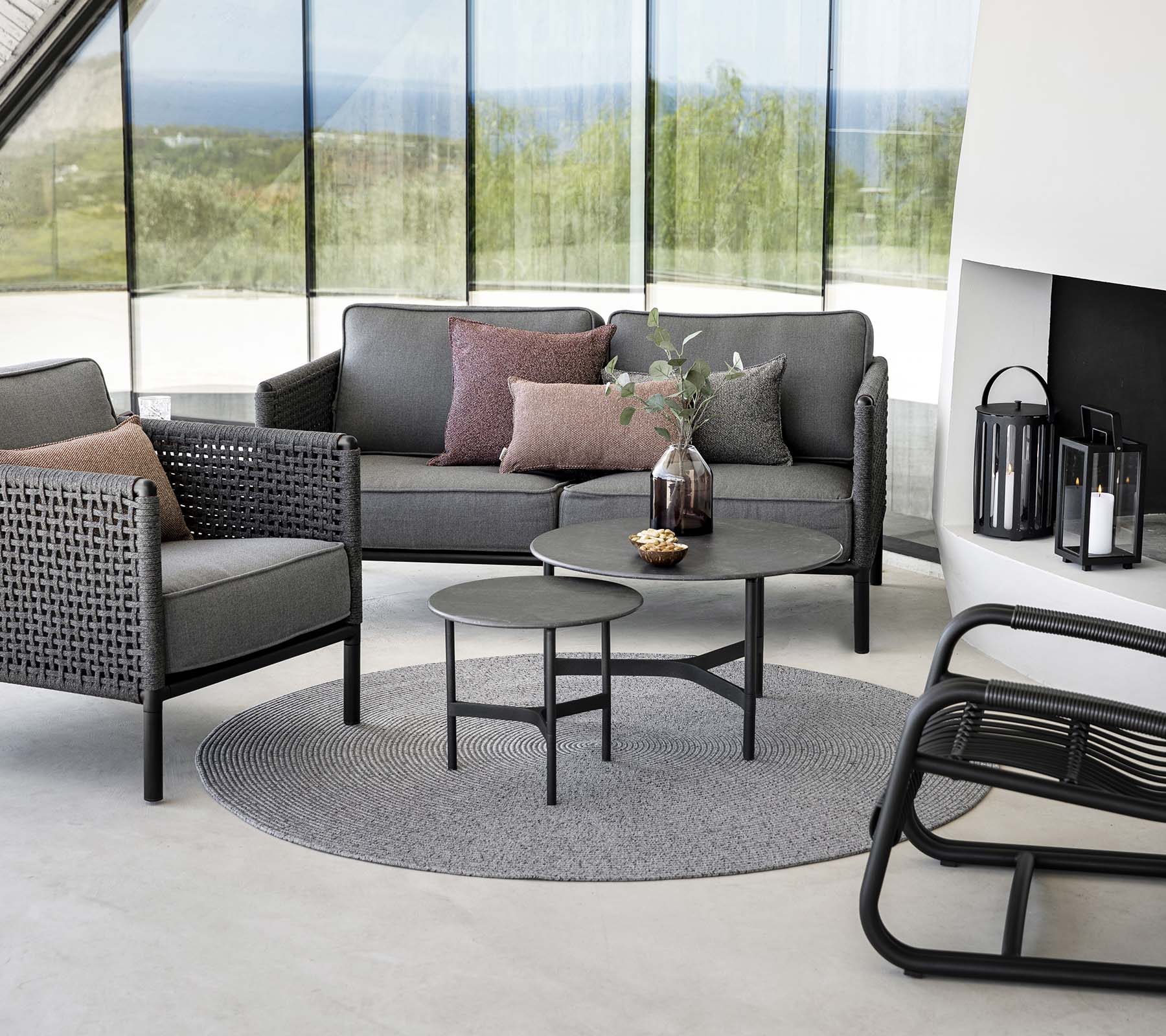 Stijlvolle zithoek met een grijze bank met kussens, een ronde salontafel en accentstoelen op een rond tapijt. Natuurlijk licht verbetert de sfeer.