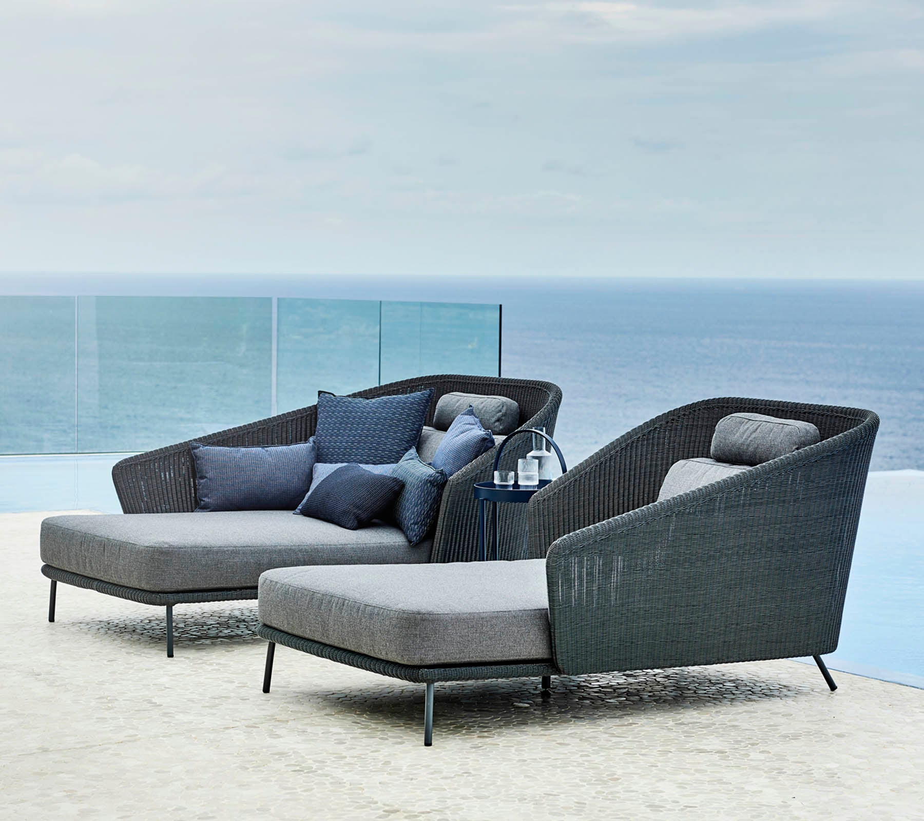 Twee stijlvolle loungestoelen met kussens en een klein bijzettafel met uitzicht op een serene oceaan.
