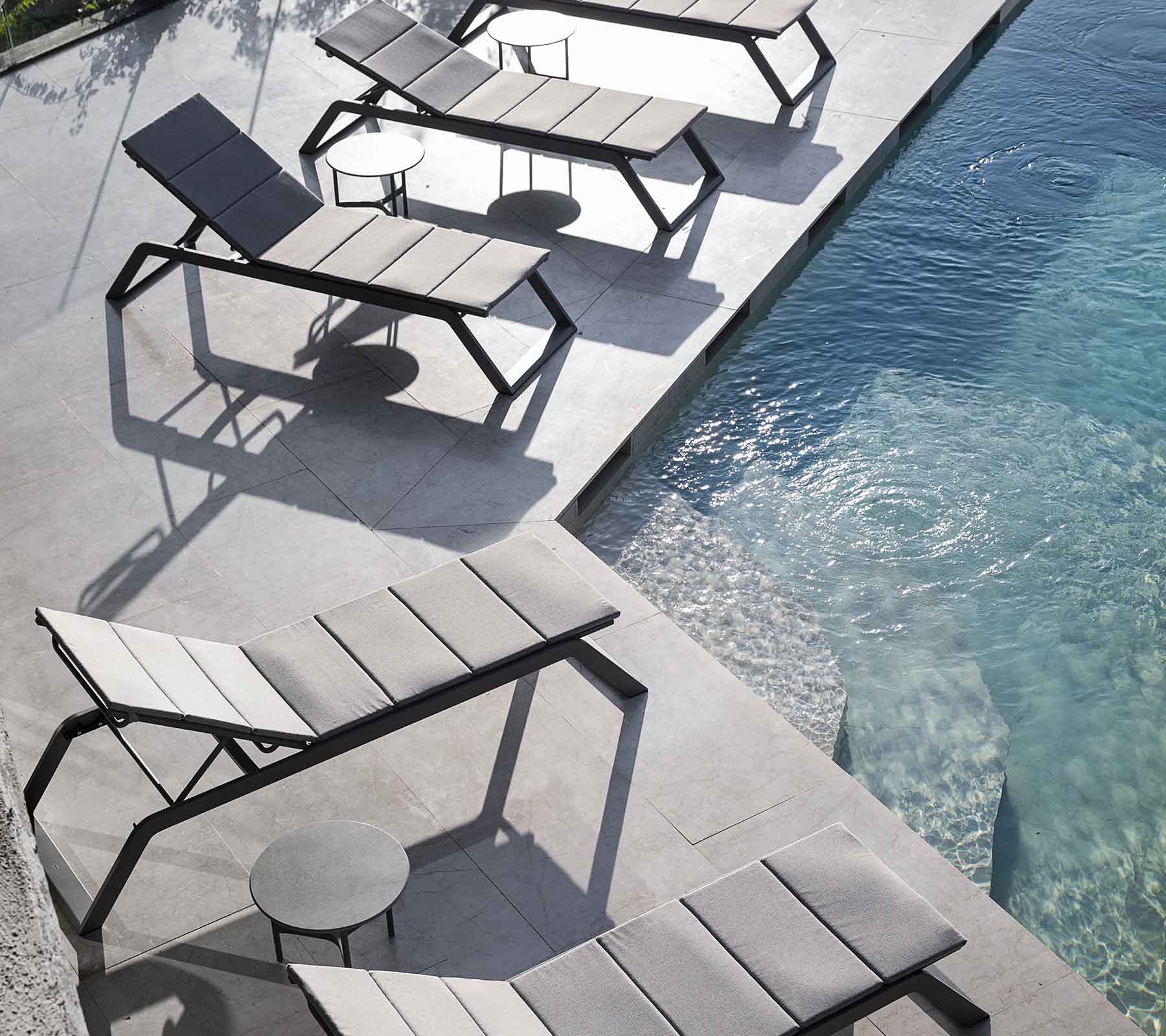 Een rij moderne lounge stoelen met een klein zijtafeltje naast een zwembad, dat schaduwen op het terras werpt.