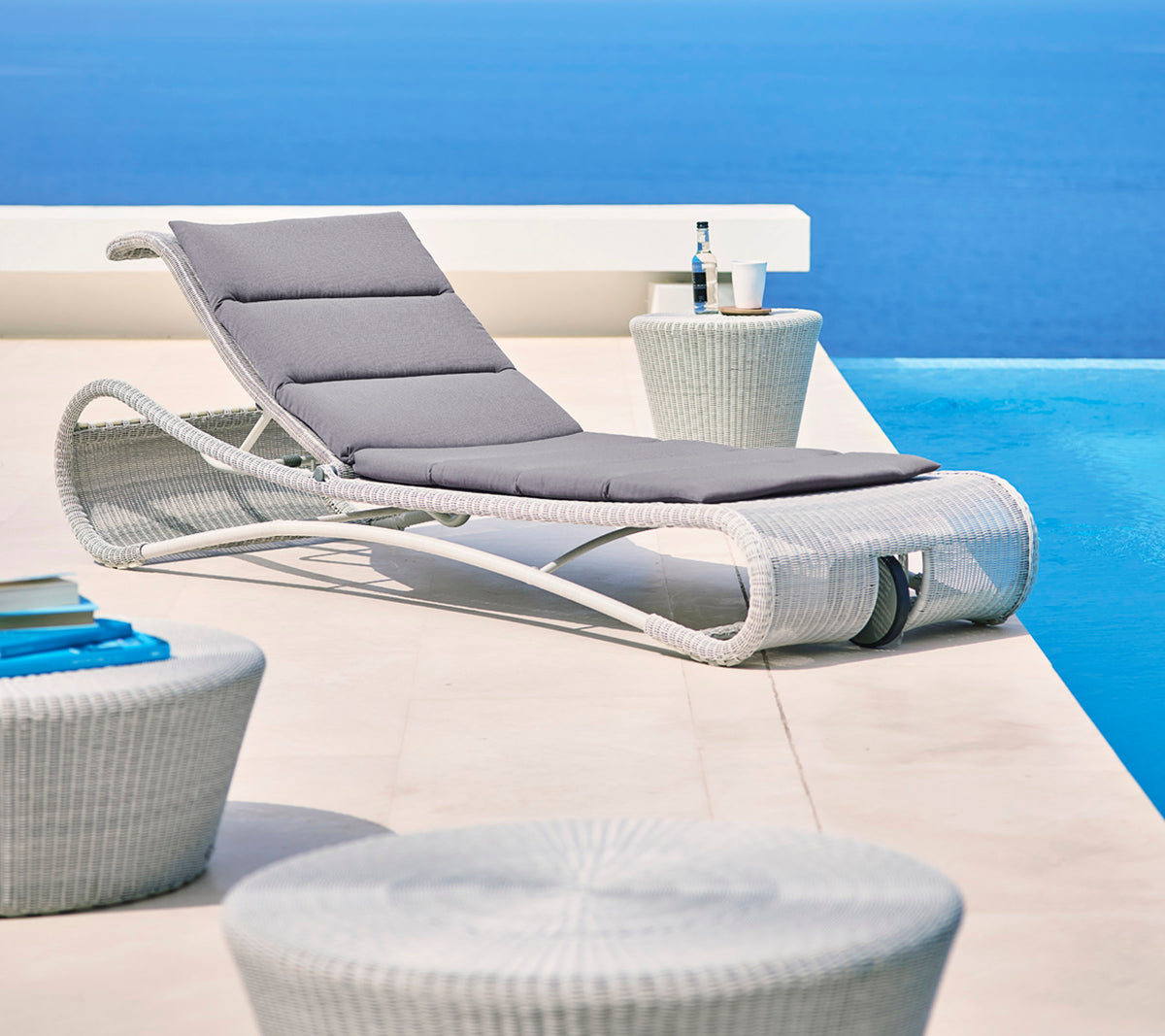 Stijlvolle loungechair met grijze kussen naast moderne sidetafels, tegen een serene blauwe achtergrond. Perfect voor ontspanning.