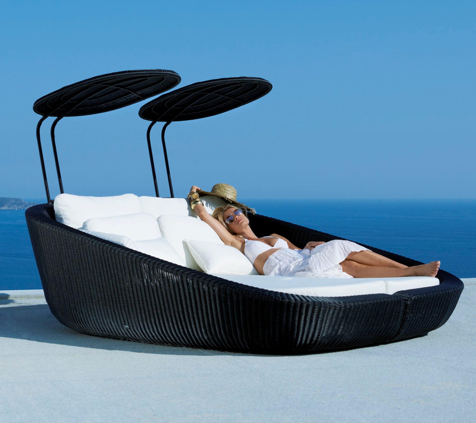 Een stijlvolle loungebed voor buiten met een slank ontwerp, voorzien van zachte kussens en verstelbare luifels, tegen een schilderachtige oceaanscène.