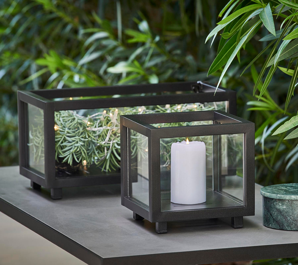 Twee stijlvolle lantaarns met glazen panelen, de ene met een witte kaars en de andere met groen, geplaatst op een tafel te midden van weelderig loof.
