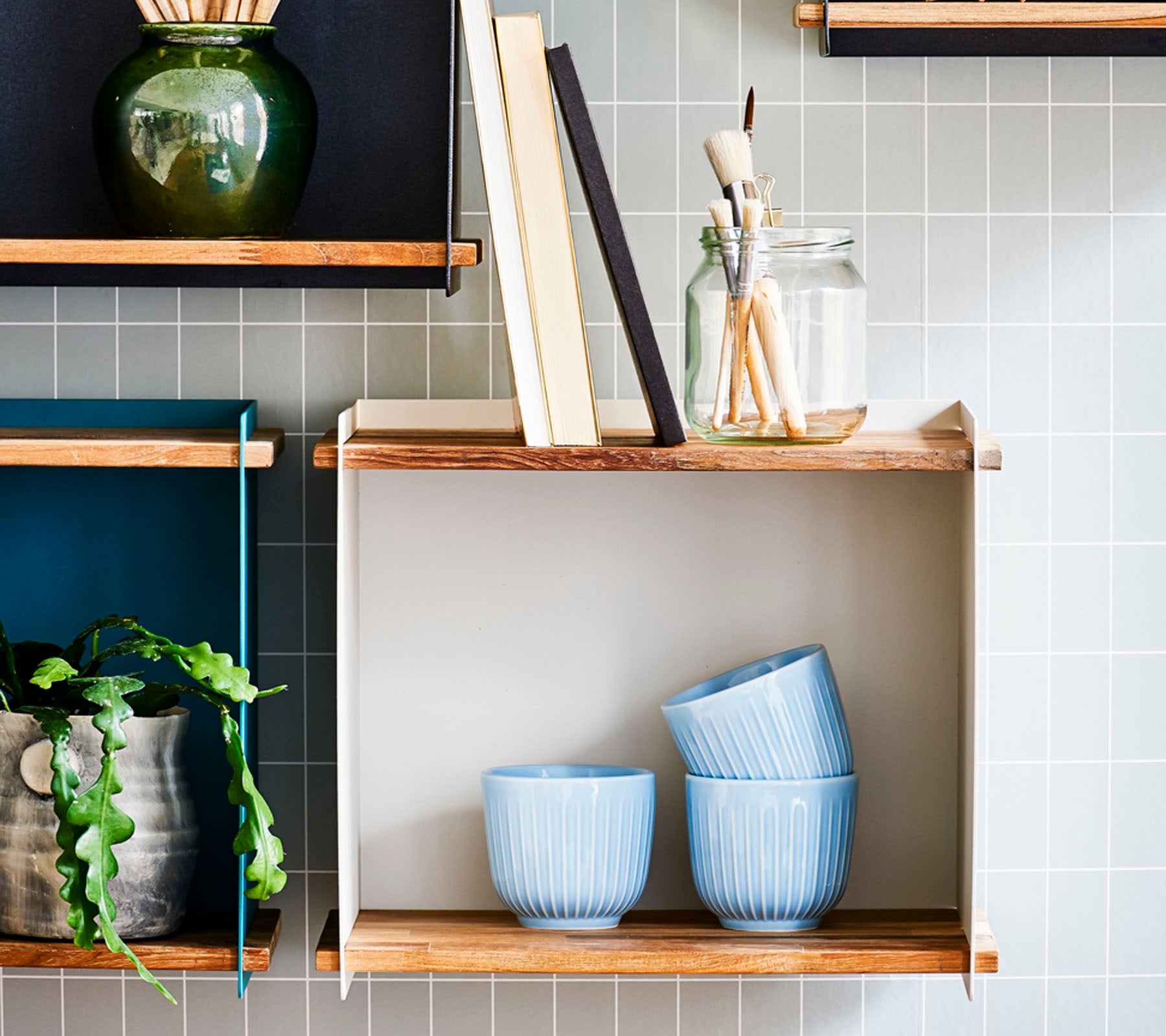 Planken met decoratieve items, waaronder blauwe kommen, boeken, een pot met penselen en een kamerplant tegen een betegelde achtergrond.