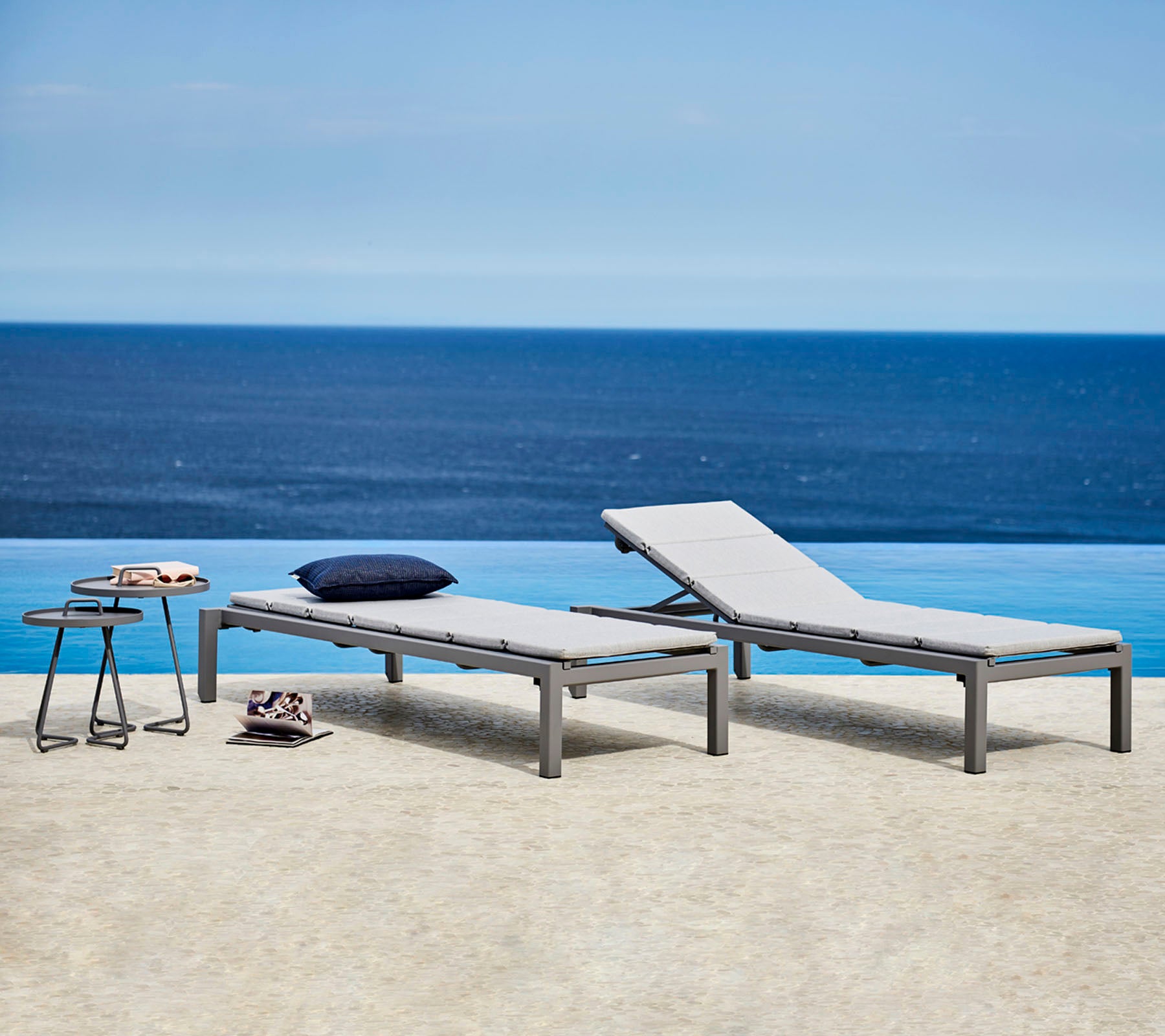 Twee stijlvolle loungestoelen met grijze kussens staan naast een klein tafeltje. De achtergrond heeft een serene uitzichten op de oceaan.