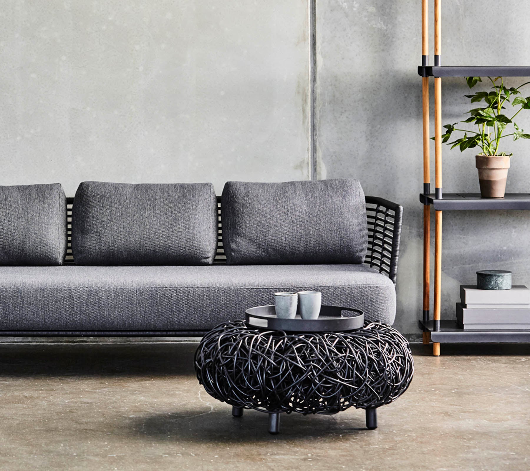 Een moderne grijze bank met kussens, gecombineerd met een rond zwart geweven salontafel en een minimalistische plank die planten en decoratie toont.