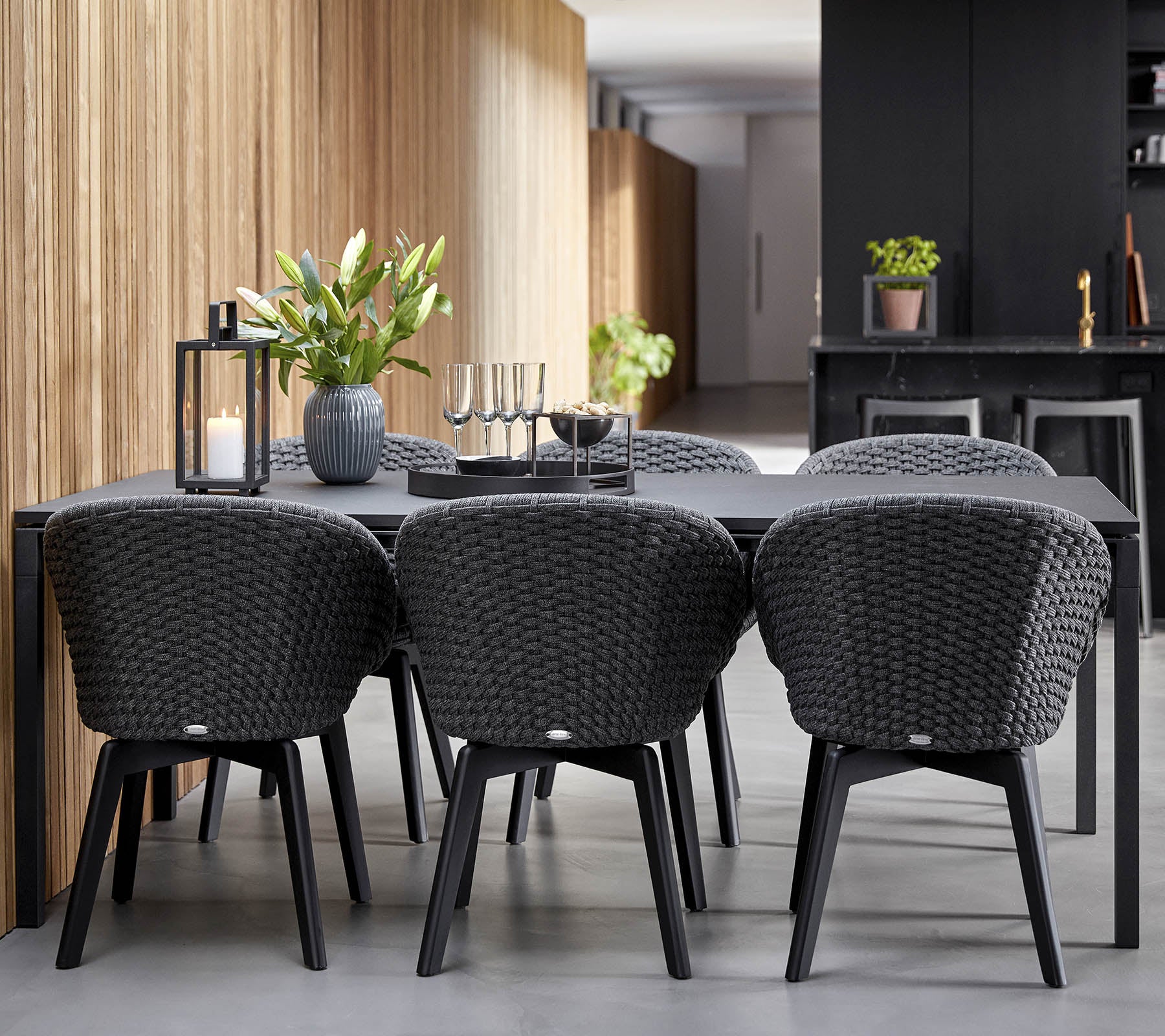 Een moderne eetopstelling met een slank zwart tafel omringd door stijlvolle, geweven stoelen, aangevuld met decoratieve planten en kaarsen.