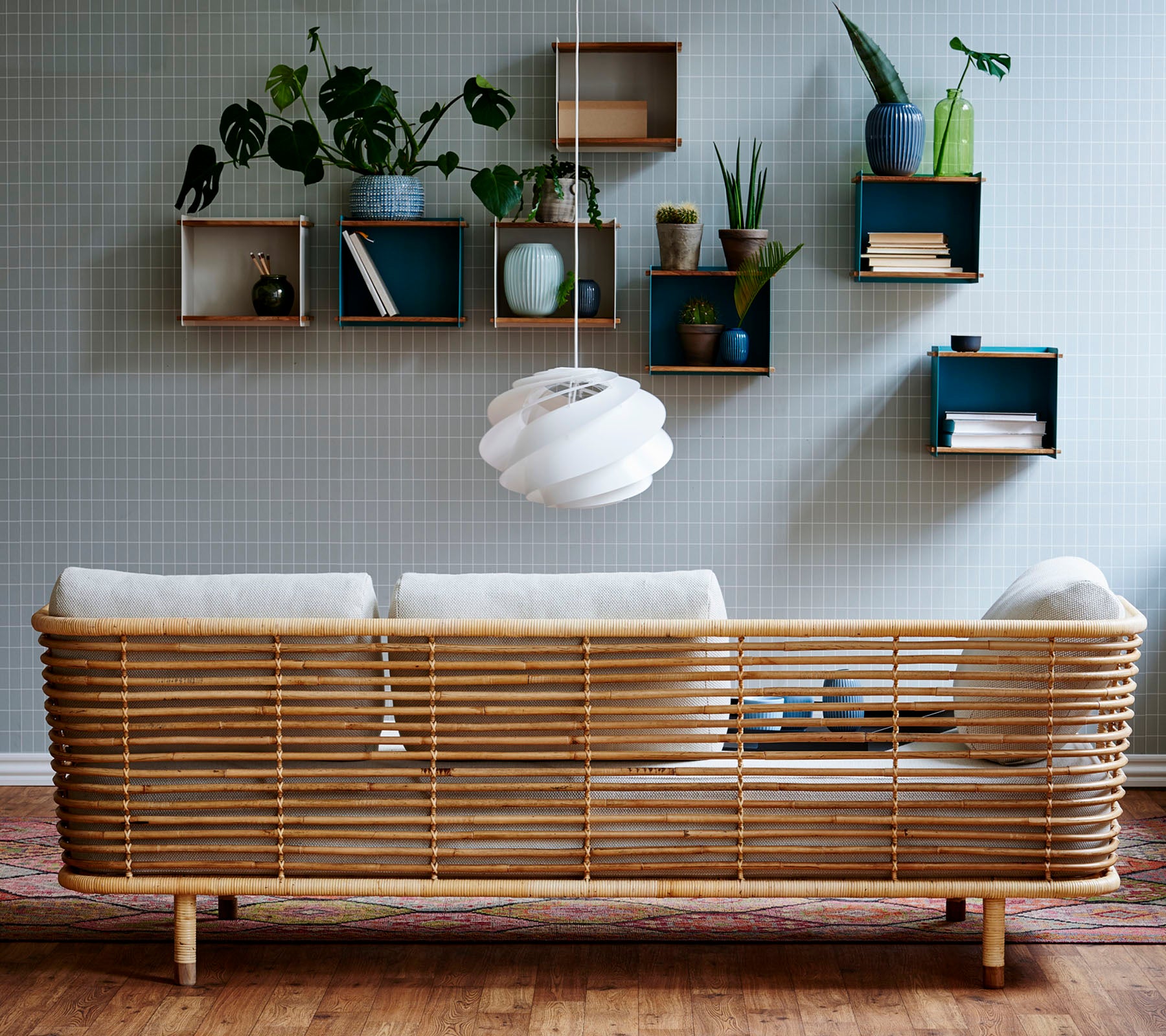Een stijlvolle rieten bank met lichte kussens tegen een gestructureerde muur, aangevuld met wandplanken die planten en decoratieve items tonen.