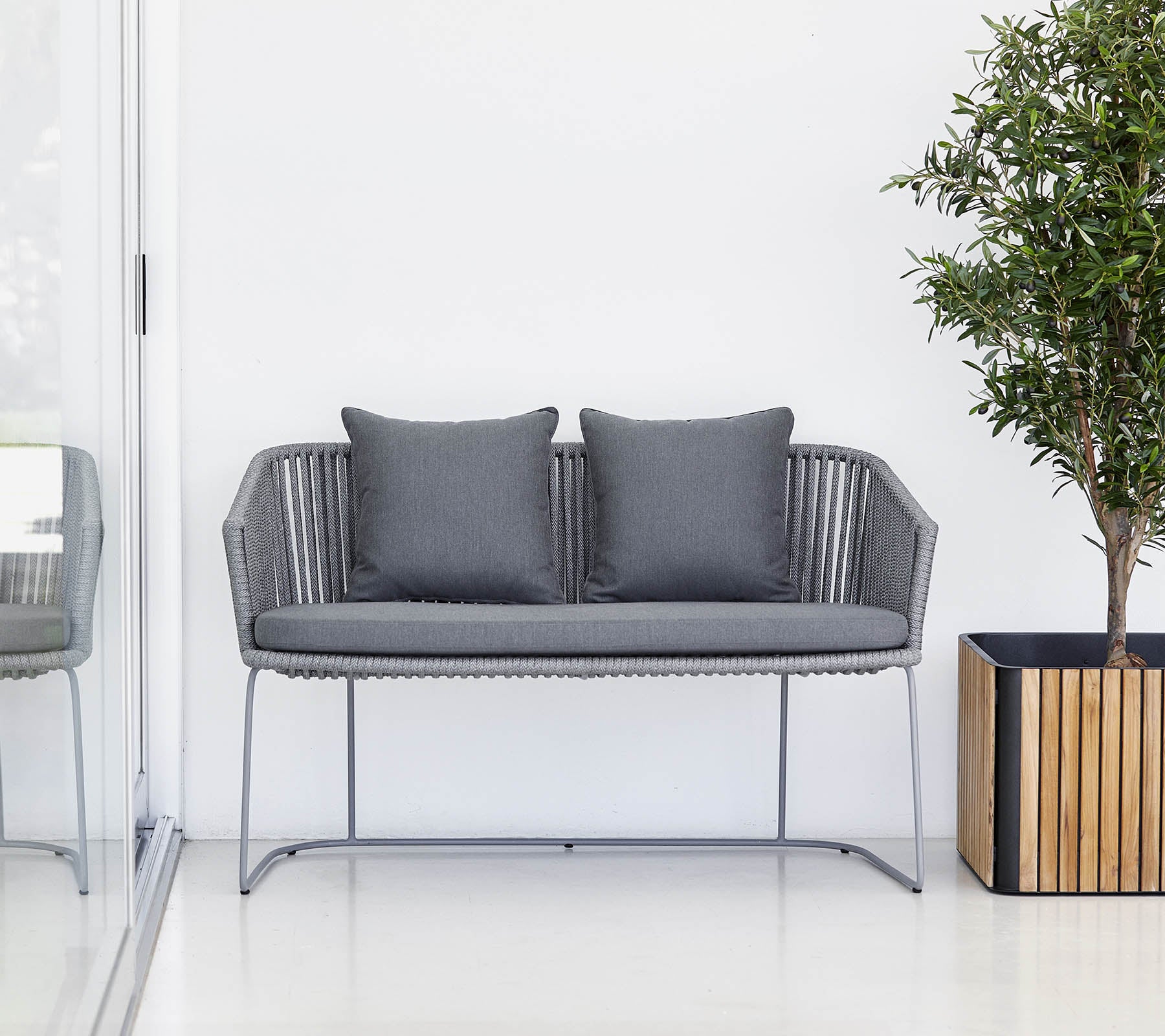 Een modern grijs tweezitsbank met kussens, vergezeld van een kamerplant en een houten plantenbak.