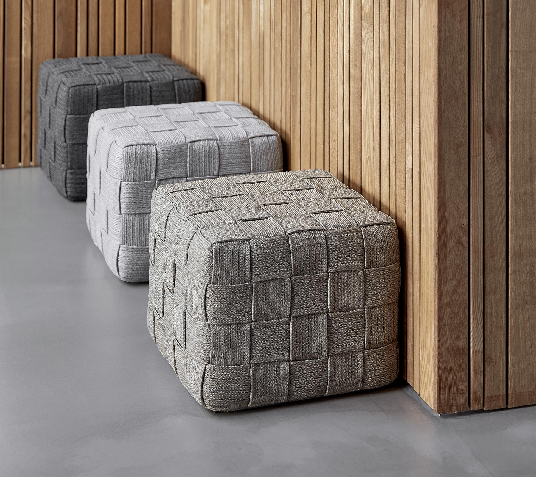 Drie textured, vierkante ottomanen in verschillende grijstinten zijn tegen een houten muur geplaatst, waardoor ze een moderne toets aan de setting geven.