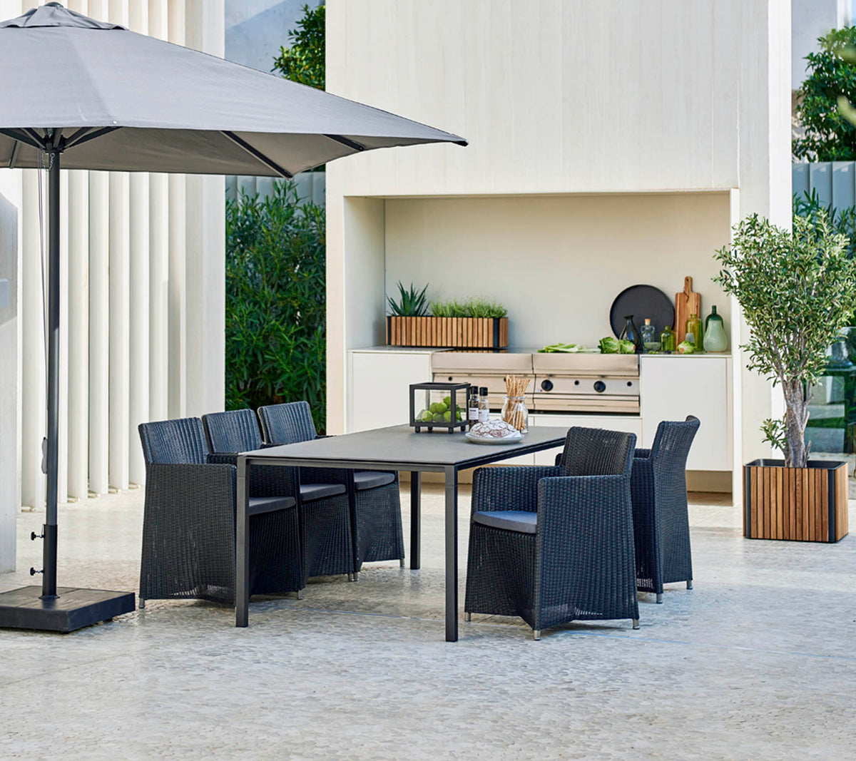 Moderne buitentafelset met een grote tafel, zes stoelen, een stijlvolle parasol en een achtergrond met groen en decoratieve elementen.