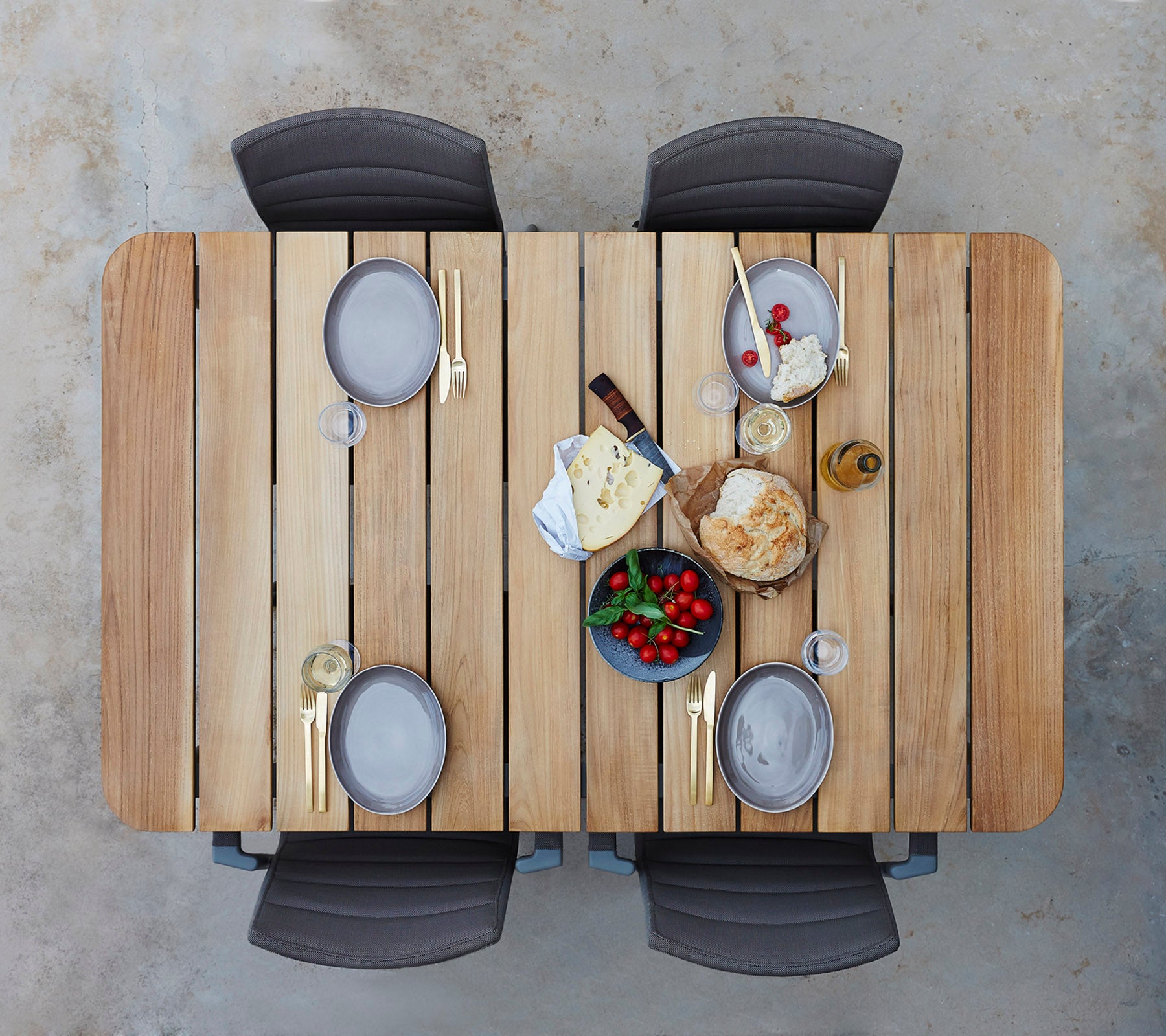 Een houten tafel gedekt met borden, glazen en een assortiment van kazen, fruit en brood, omringd door vier stoelen.
