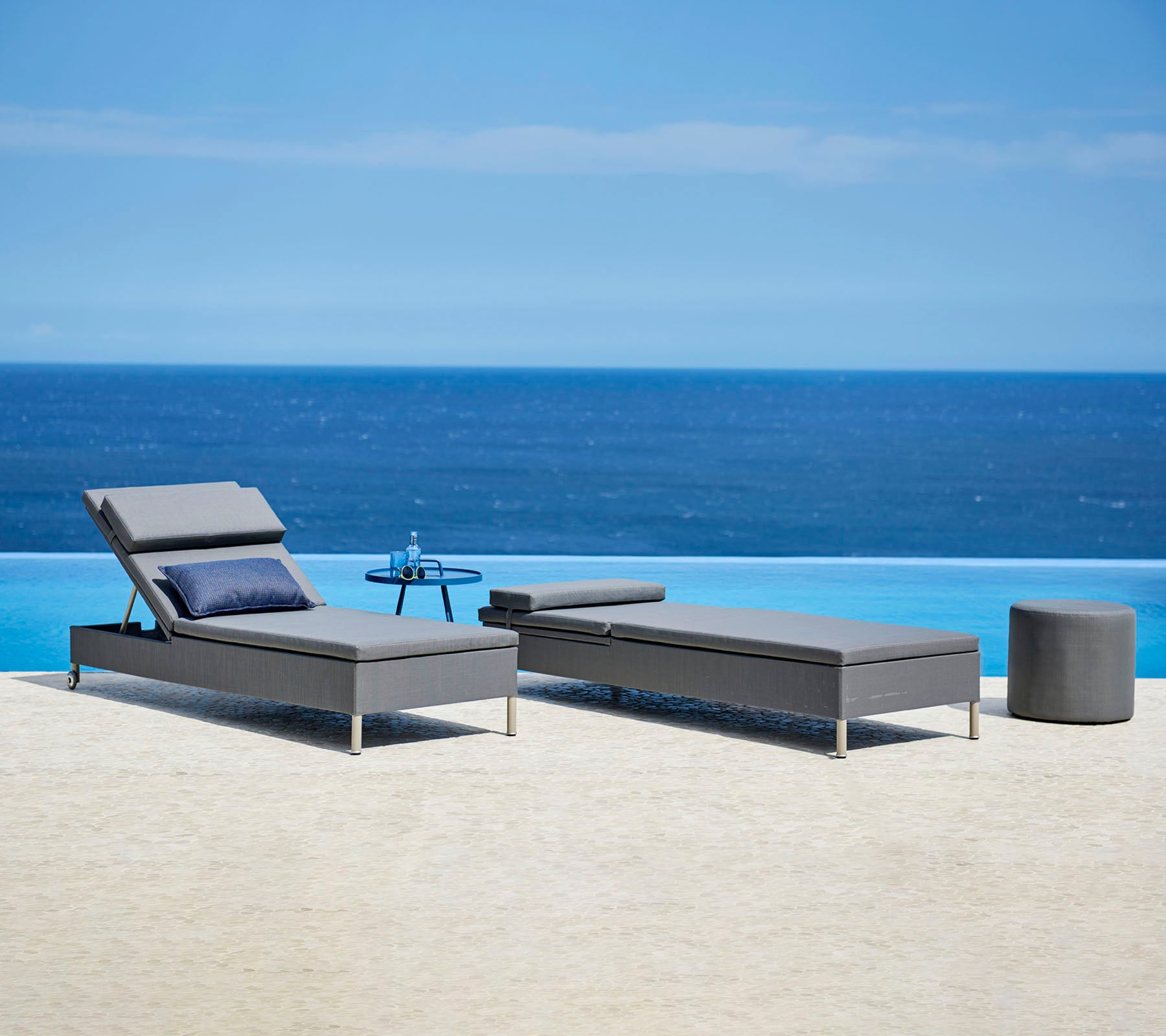 Twee stijlvolle grijze ligstoelen met kussens en een klein bijzettafeltje bij een rustig blauw waterkenmerk, perfect om te ontspannen.