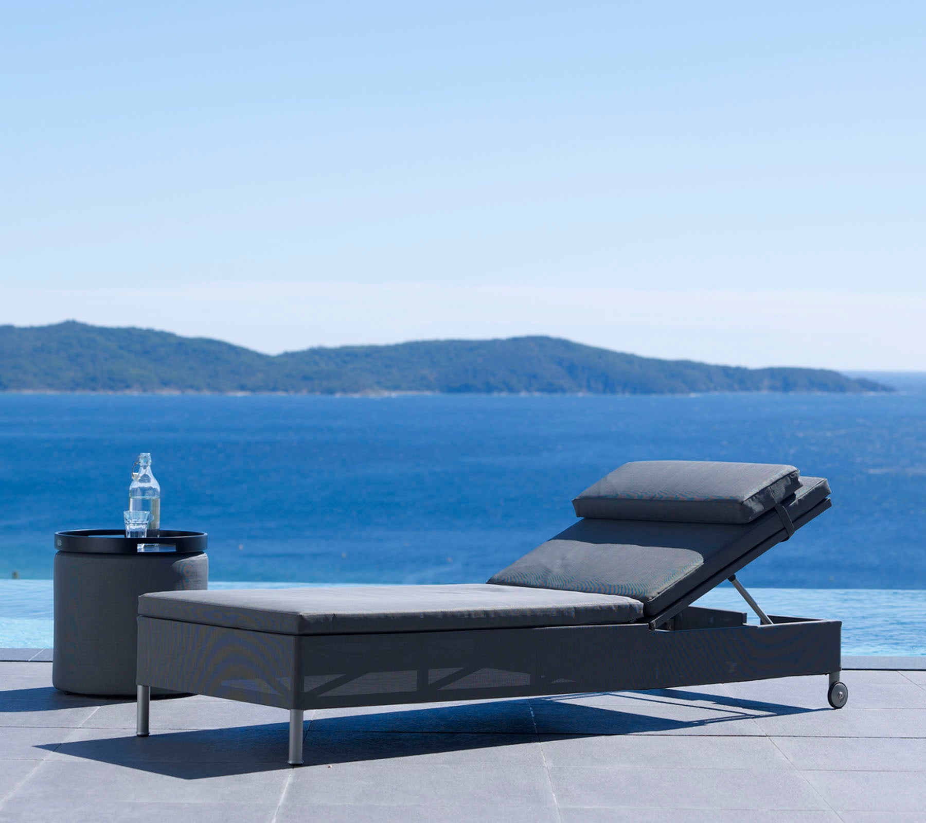 Een moderne loungestoel en bijzettafel naast een sereen waterlichaam met verre heuvels onder een heldere lucht, perfect om te ontspannen.