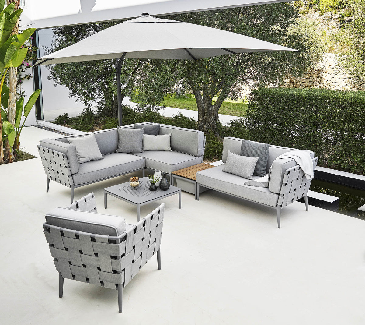 Stijlvolle buitenzitgelegenheid met een grijze hoekbank, fauteuil en salontafel onder een grote parasol.
