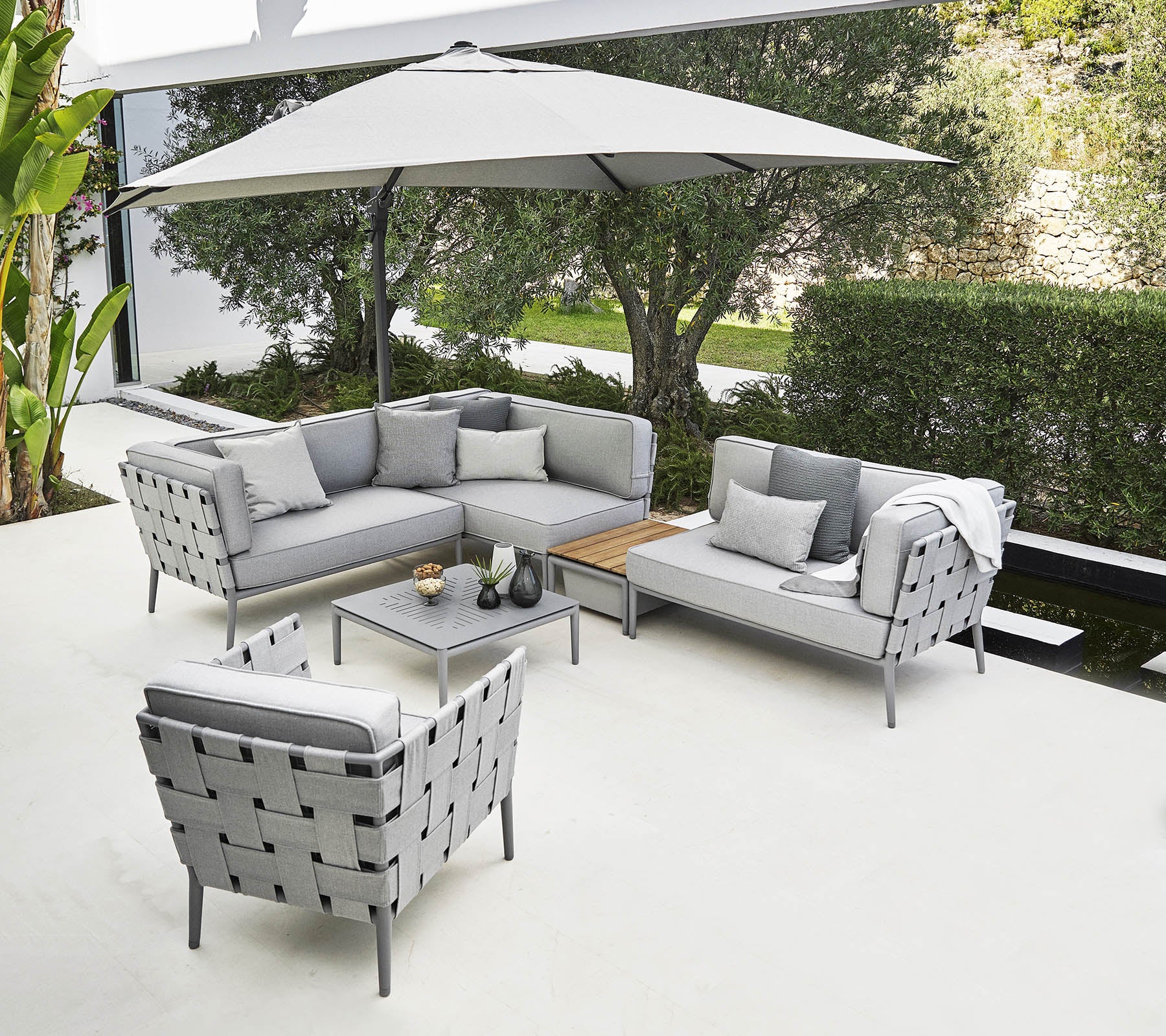 Stijlvolle buitenzitgelegenheid met een grijze hoekbank, fauteuil en salontafel onder een grote parasol.
