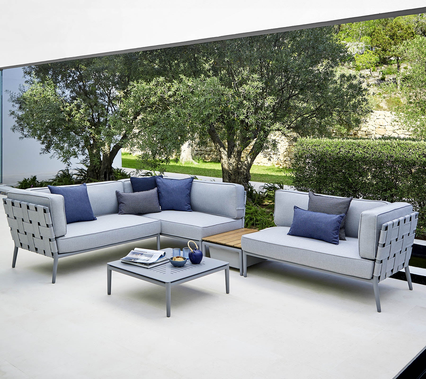 Een moderne buitenzitopstelling met lichtgrijze sofa's met blauwe kussens en een bijpassende salontafel, tegen een weelderige achtergrond.