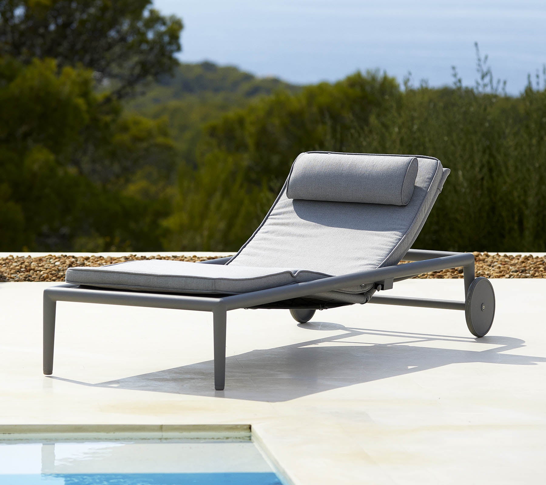 Comfortabele loungechair met een grijze kussen en rugleuning, voorzien van wielen voor gemakkelijke mobiliteit, naast een zwembad en weelderig groen op de achtergrond.