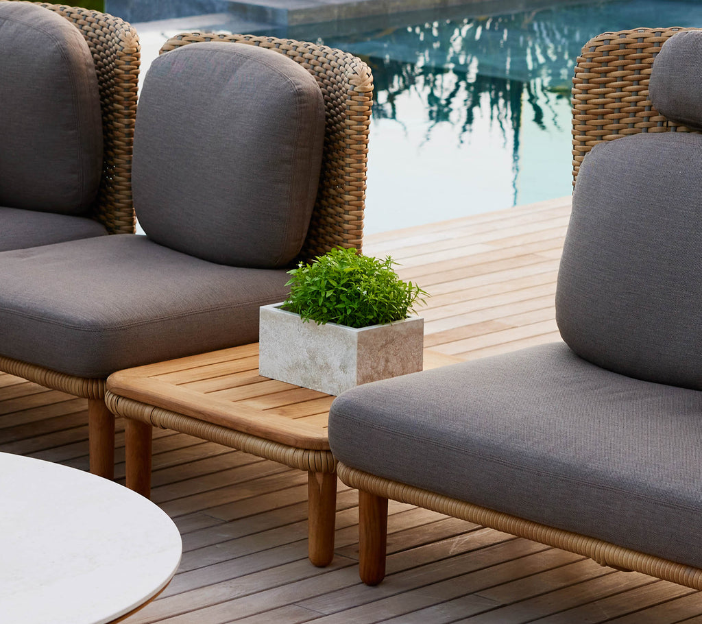 Een stijlvol Arch salontafel met een teak tafelblad, geflankeerd door loungerstoelen en een kleine potplant, wat een chique buitenomgeving versterkt.