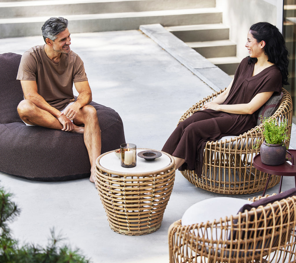 Twee personen zitten samen, de een op een gezellige loungestoel en de ander op een ronde kuipstoel, met een klein rieten salontafeltje tussen hen in.