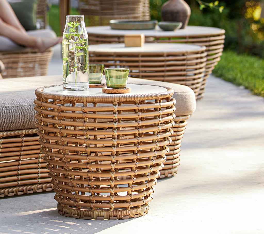 Klein mand koffie tafel met een geweven ontwerp en een glazen blad, vergezeld van een waterfles en groen glas.