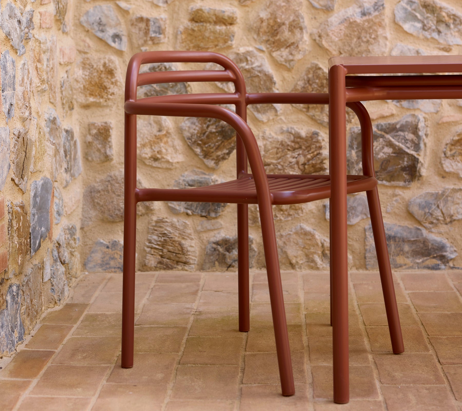 Woestijnrode Bliss-fauteuil naast een bijpassende tafel, geplaatst tegen een gestructureerde stenen muur. Een stijlvolle en moderne zitopstelling.