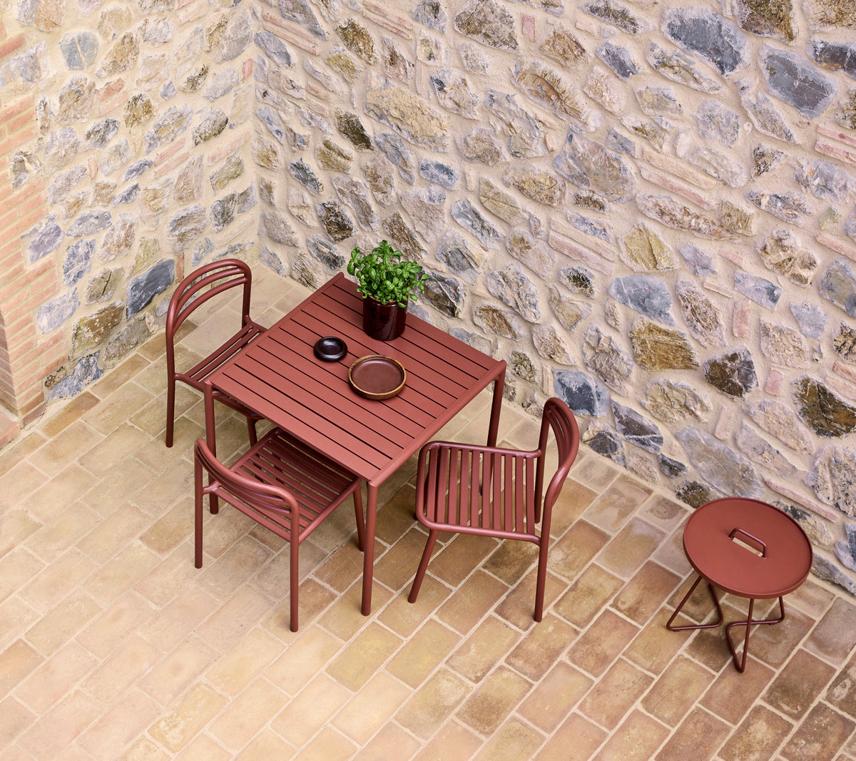 Een tafel en stoelen in een woestijnrode tint, naast een kleine ronde tafel, tegen een gestructureerde stenen muur met een potplant.
