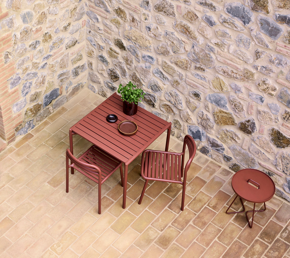 een rode café tafel gezet met twee stoelen, met een kleine plant en een bord, tegen een stenen muur.