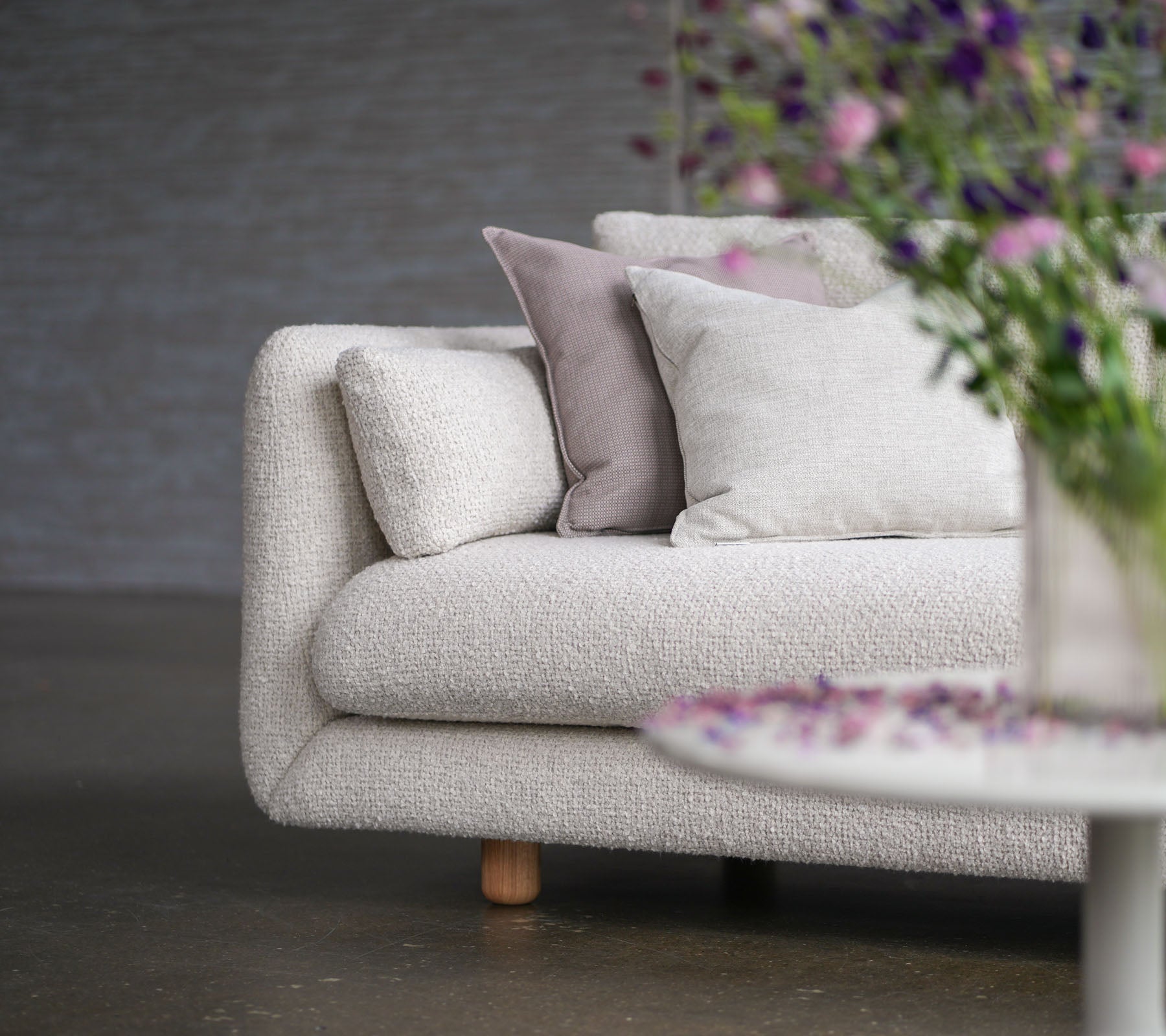 Zachte sofa met zandkleurige bekleding, decoratieve kussens, teak benen en een vaas met bloemen, die de flair en elegantie van Cane-line toont.