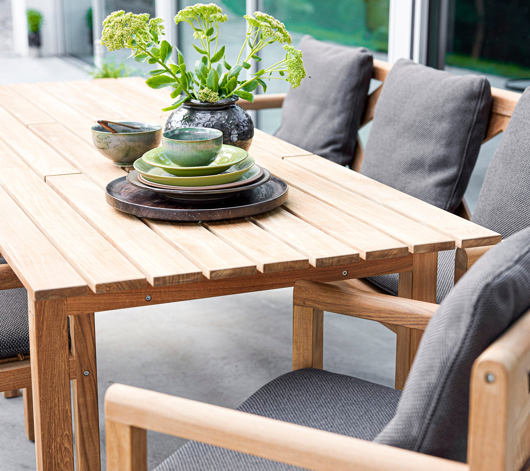 Stijlvolle houten tafelset met kussens, borden en schalen, geaccentueerd door een vaas met groen. Perfect voor uitnodigende bijeenkomsten.