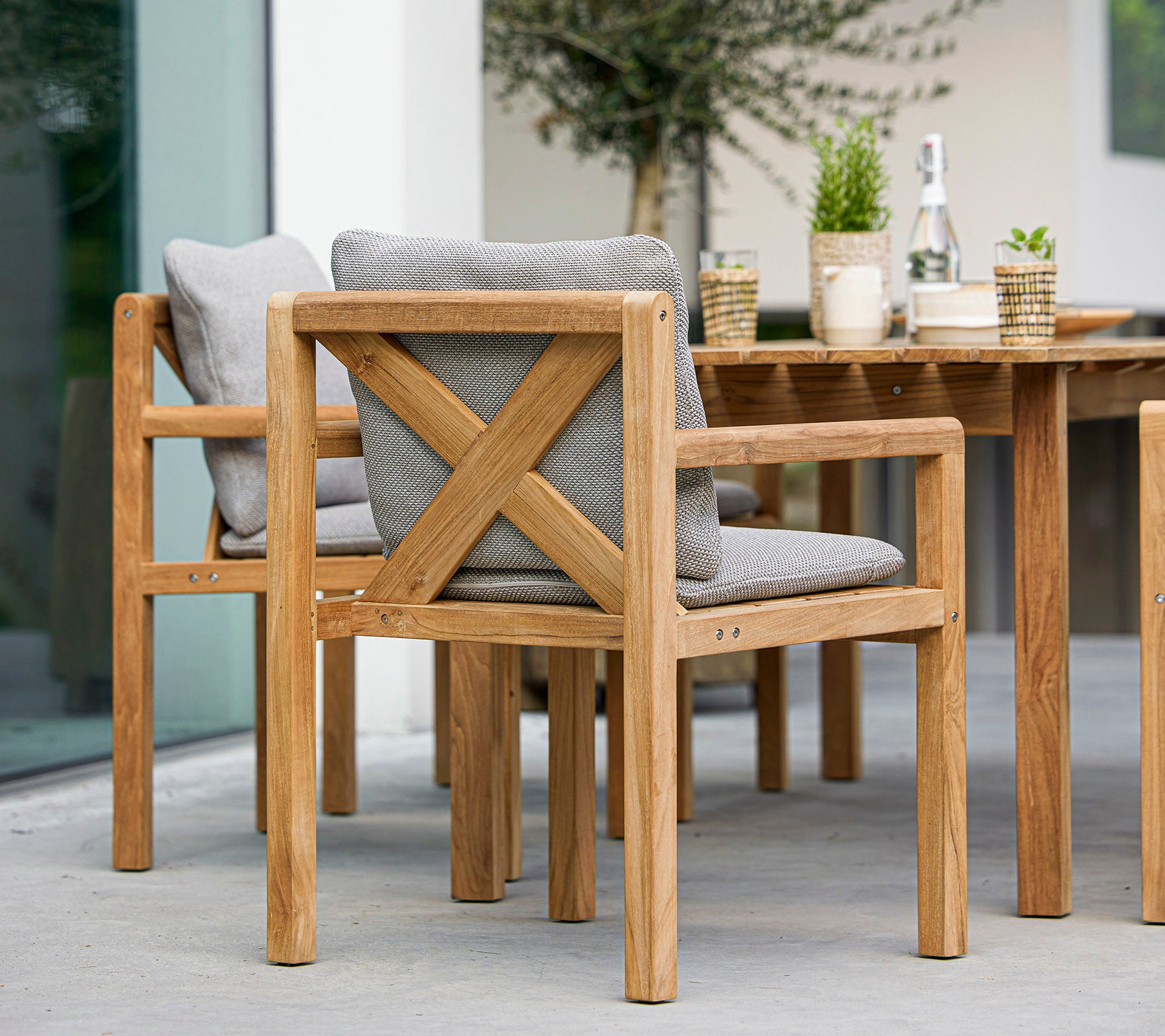 Houten stoelen met grijze kussens en een bijpassende tafel, vergezeld van decoratieve planten en een fles, ideaal voor buiten dineren.