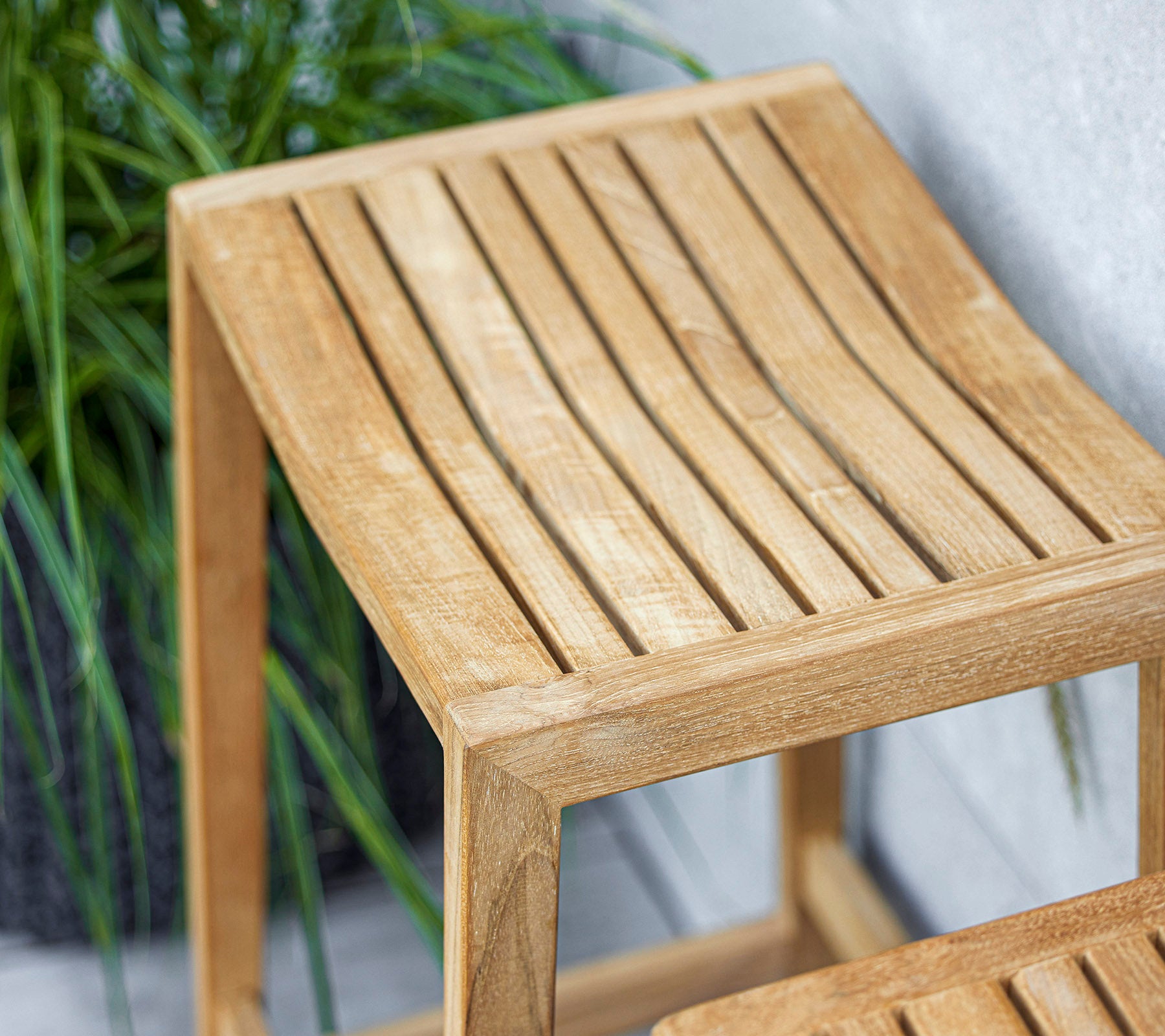 Een strak houten zijtafeltje met een lattenontwerp aan de bovenkant, met een lichte afwerking die de omliggende groenvoorziening aanvult.