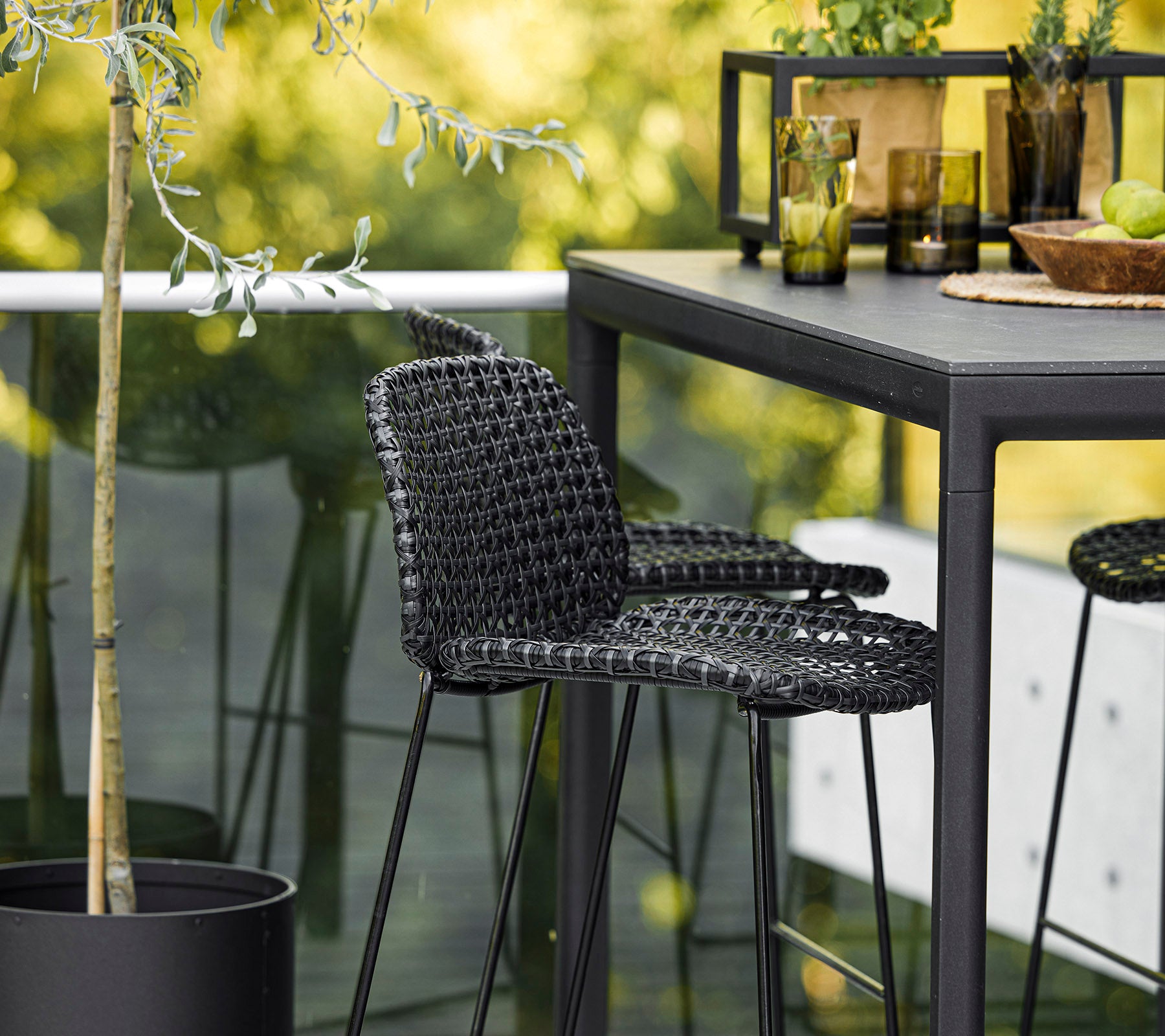 Een stijlvolle zwarte gevlochten stoel in combinatie met een strak tafeltje, omringd door decoratieve planten en glaswerk, waarmee een moderne setting wordt gecreëerd.