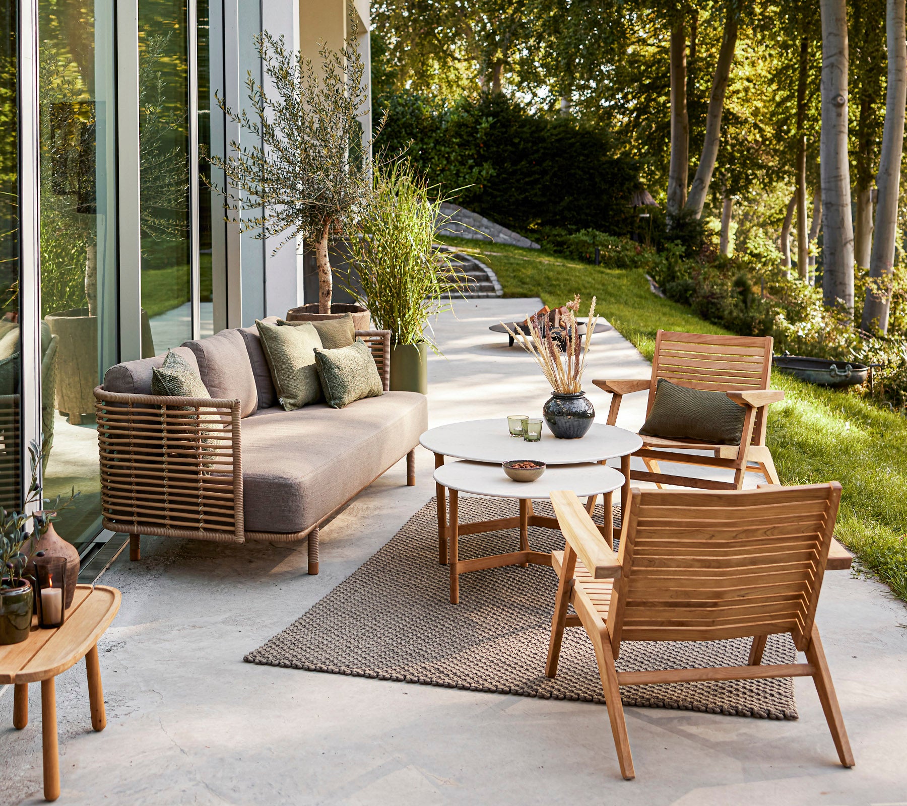 Stijlvolle buitenzitruimte met een moderne bank, houten stoelen, een ronde tafel en decoratieve planten tegen een serene achtergrond.