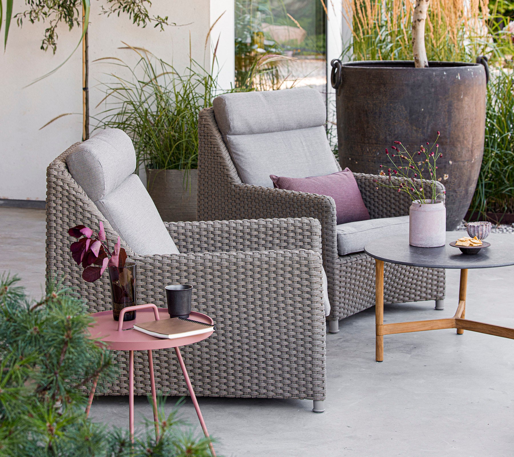 Twee comfortabele grijze stoelen met kussens, een ronde tafel met snacks en drankjes, omringd door groen en decoratie. Perfect voor ontspanning.