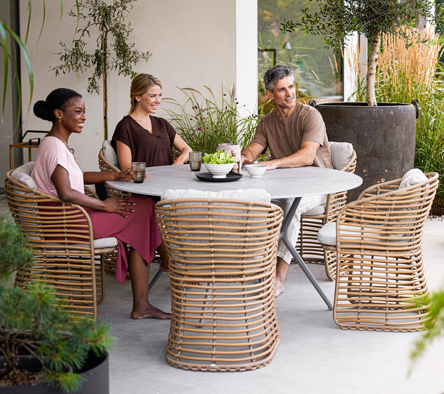 Vier mensen genieten van drankjes rond een modern rond tafel, met stijlvolle rieten stoelen en groen in een serene setting.