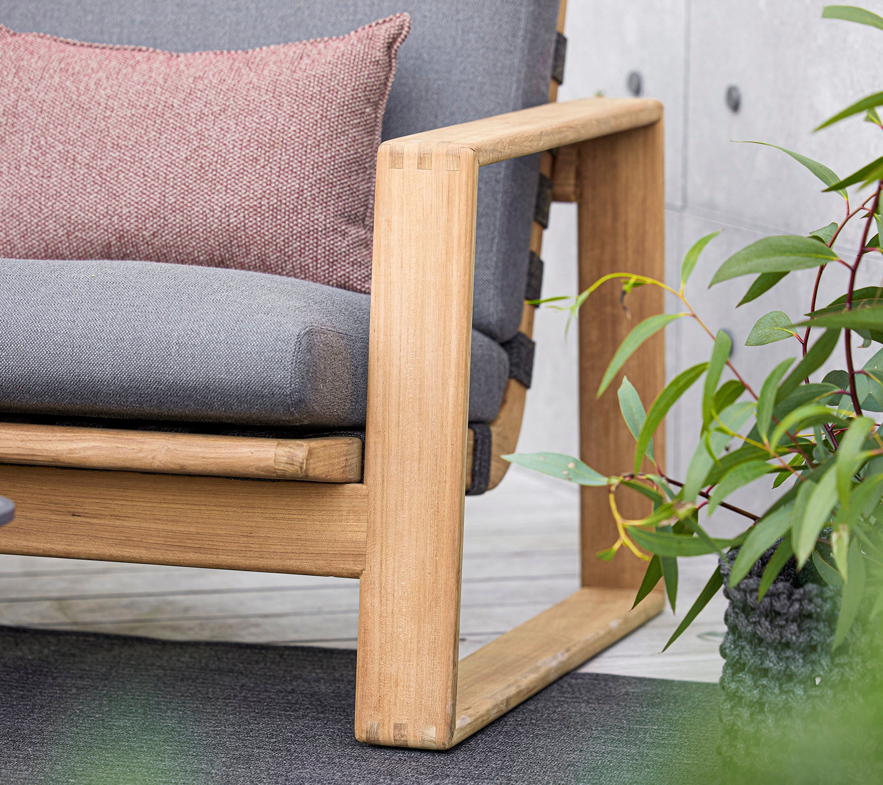 een modern bank met een houten frame, voorzien van grijze kussens en een decoratief kussen, aangevuld met weelderige groene planten in de buurt.