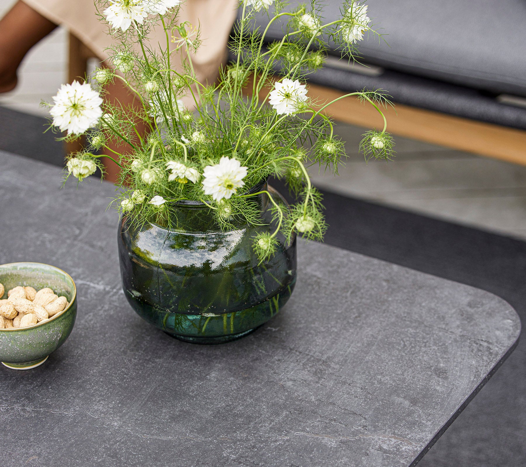 Een modern tafelblad met een ronde glazen vaas met witte bloemen en een kleine schaal met noten. Het oppervlak is glad en gestructureerd.